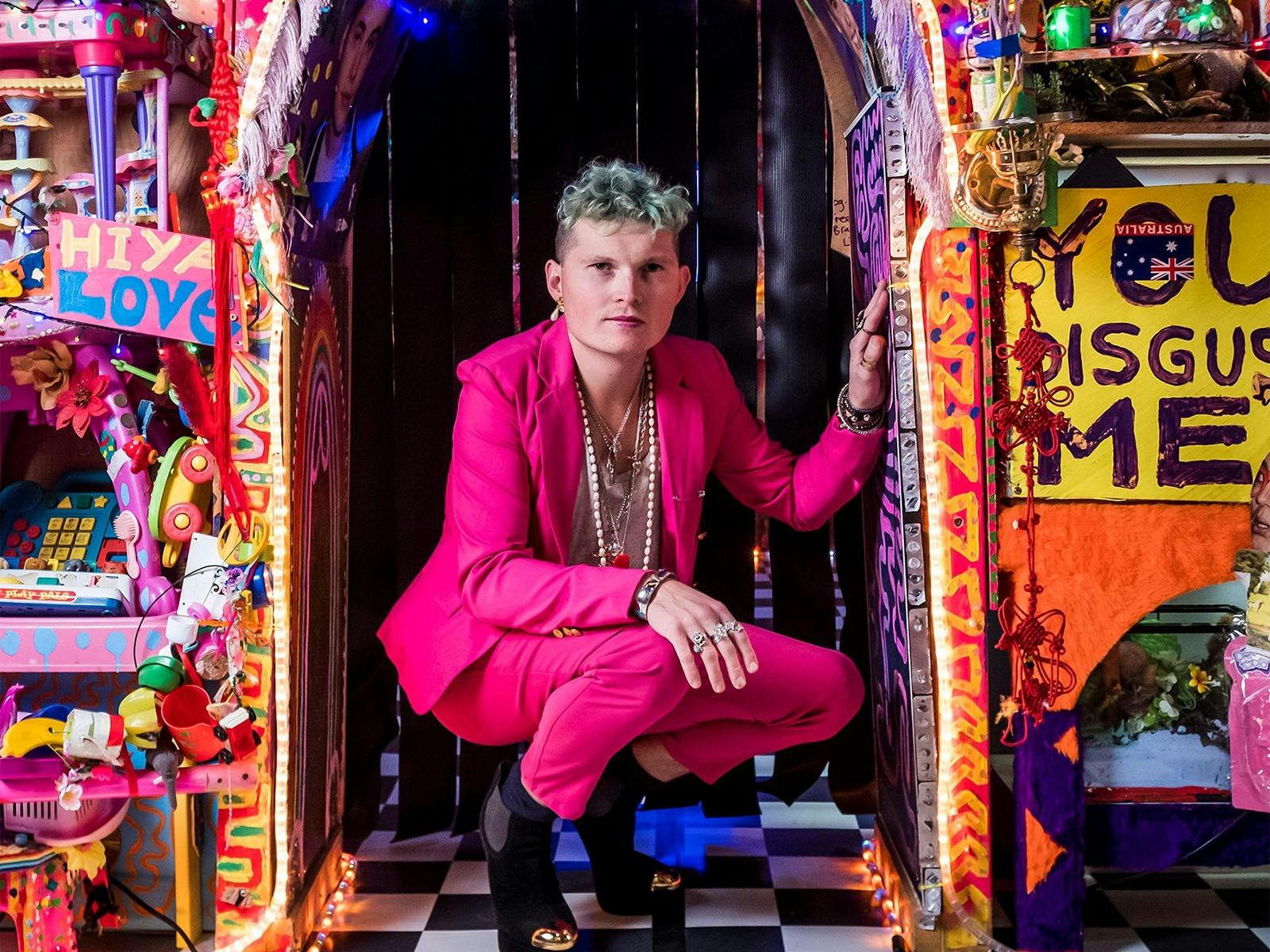 An adult wearing a pink suit poses on the ground in a colourful, decorated room.