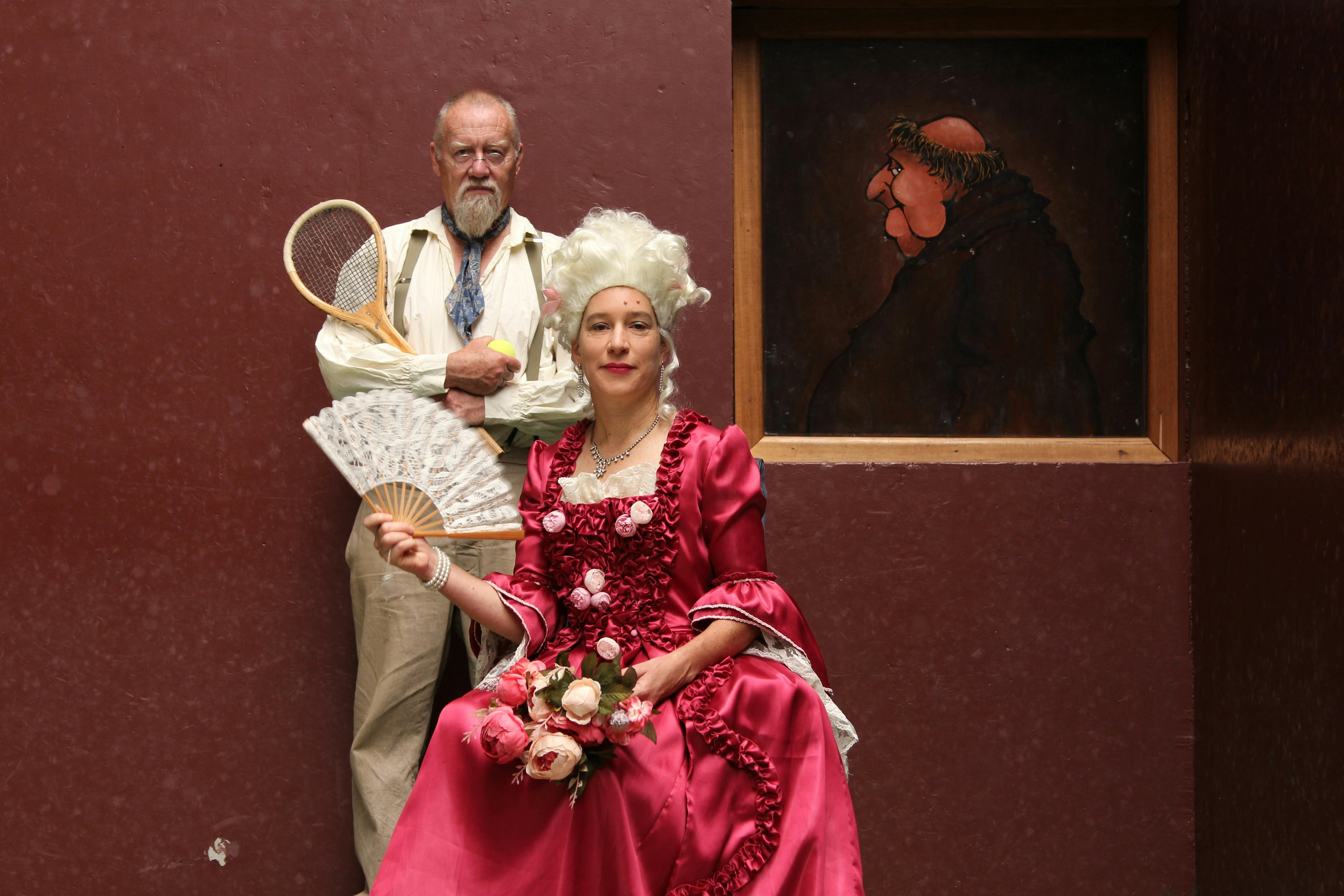 A man in 19th century dress stands behind a seated woman in 18th century dress near a painting.