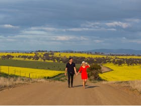 two people walking on dirt road with canola paddocks in the background