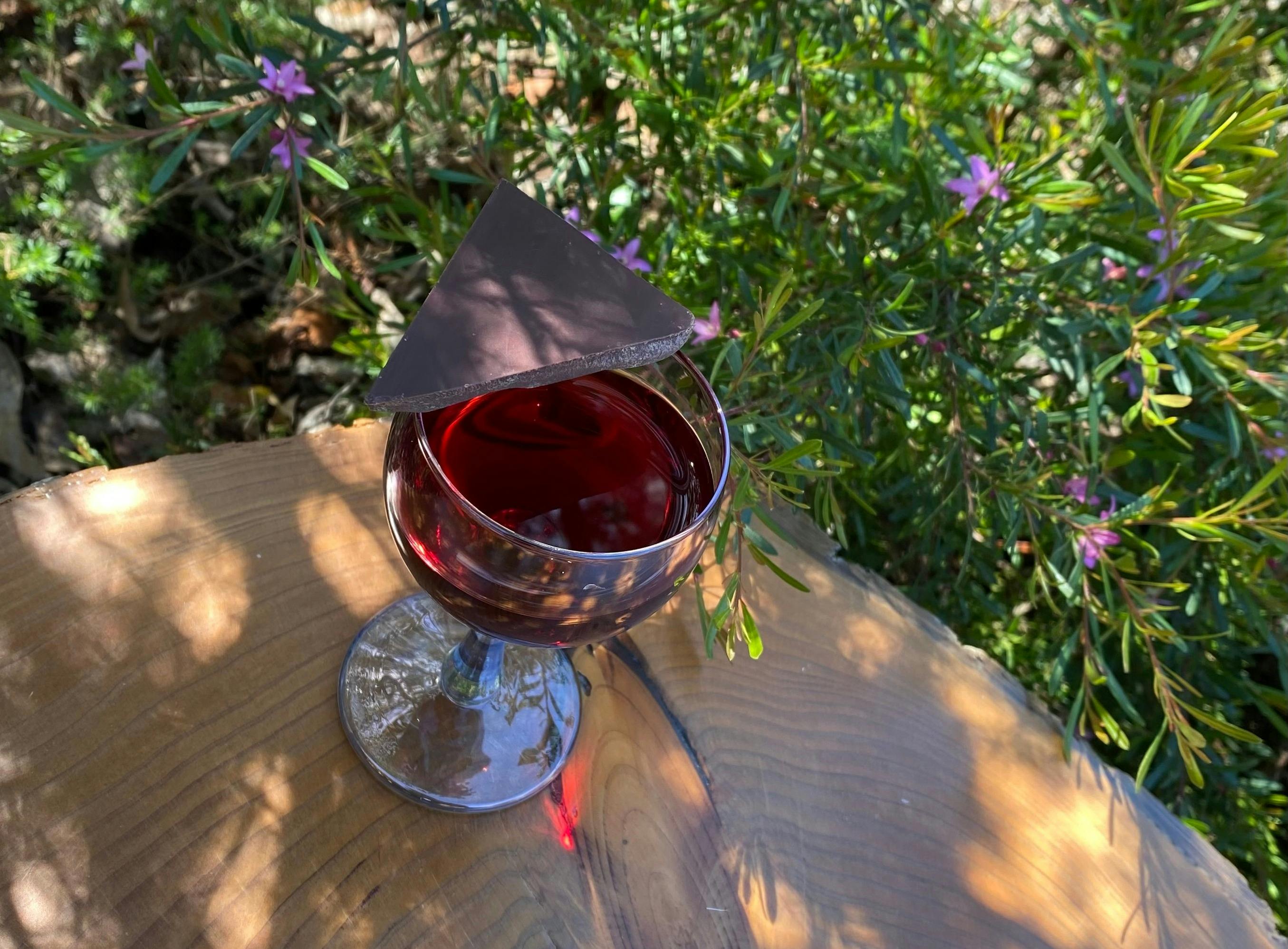 A glass of wine with dark chocolate balanced over half of the glass atop a wooden table in a garden