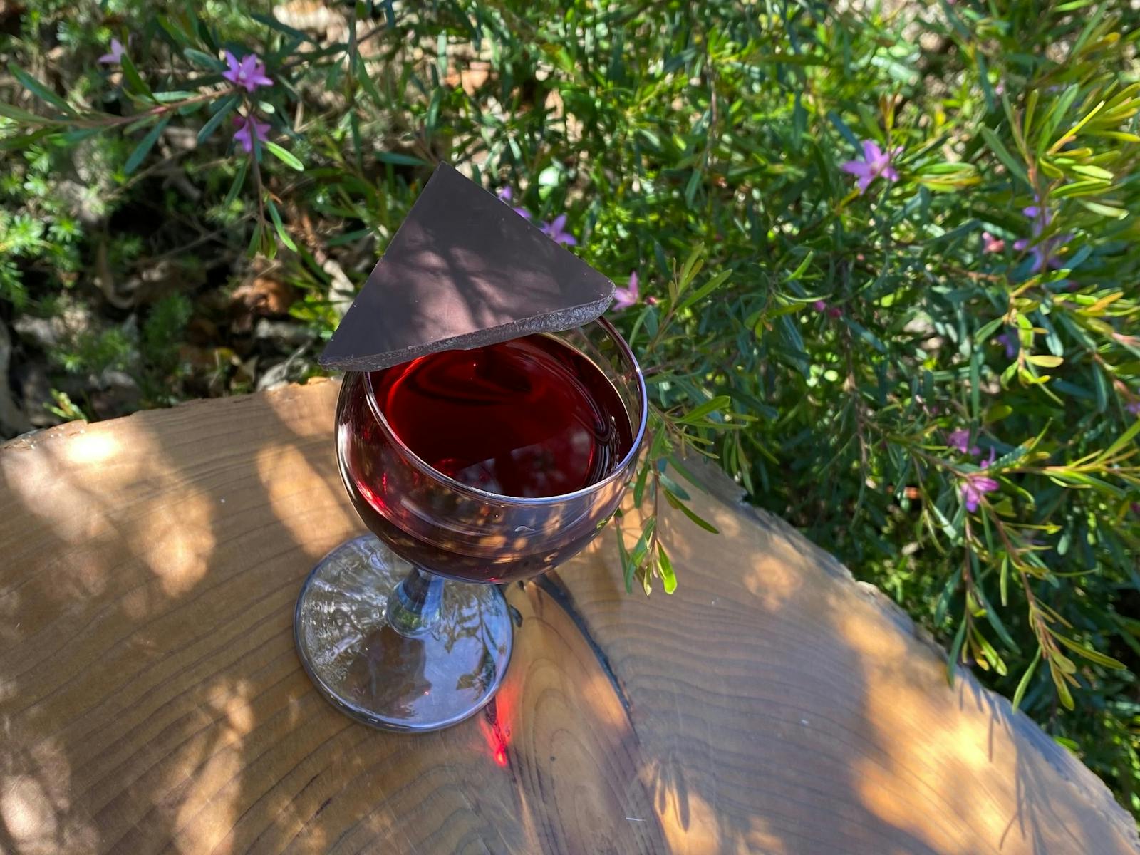 A glass of wine with dark chocolate balanced over half of the glass atop a wooden table in a garden