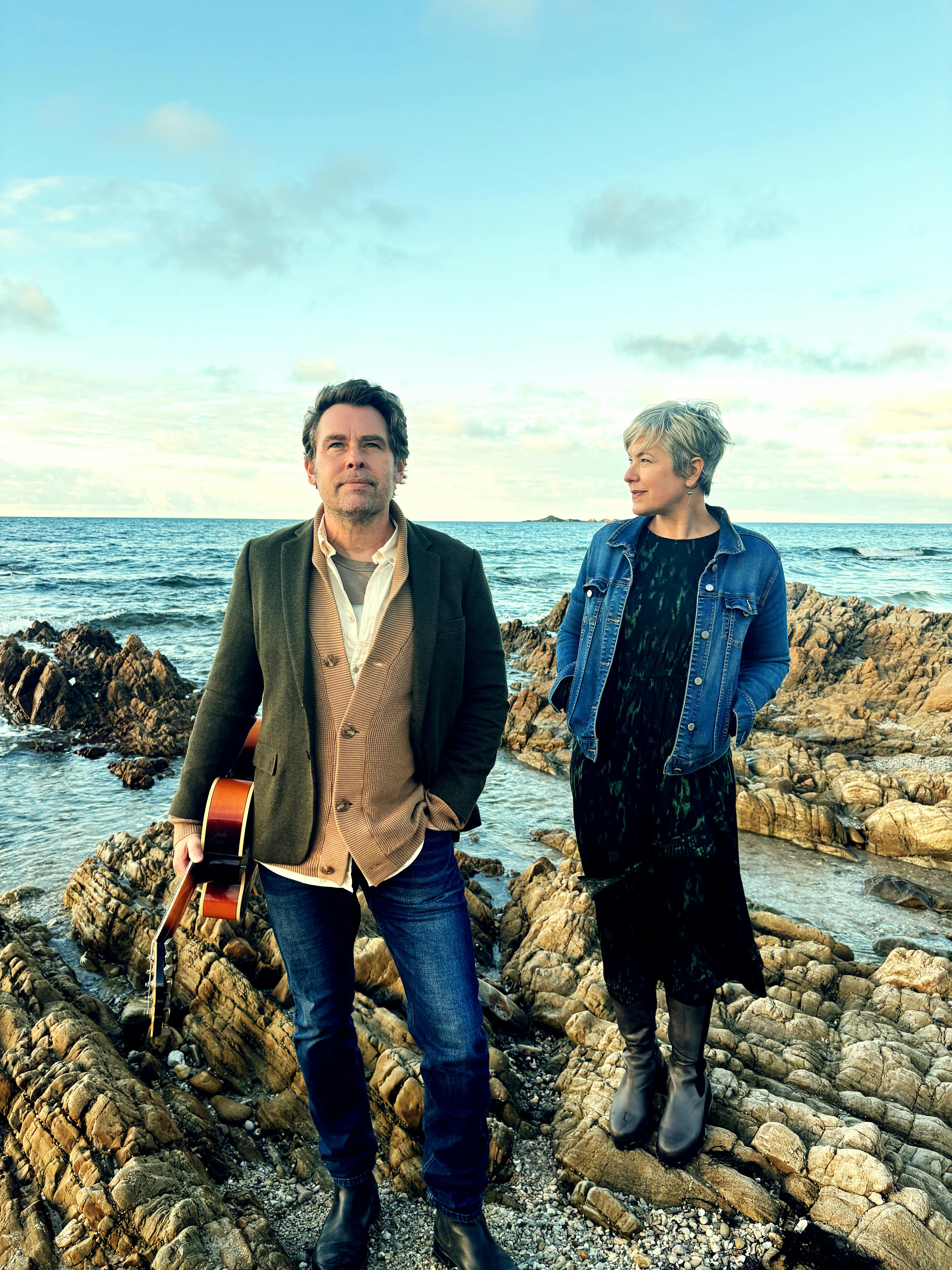 A man and a woman stand on rocks at the beach. the man holds a guitar.