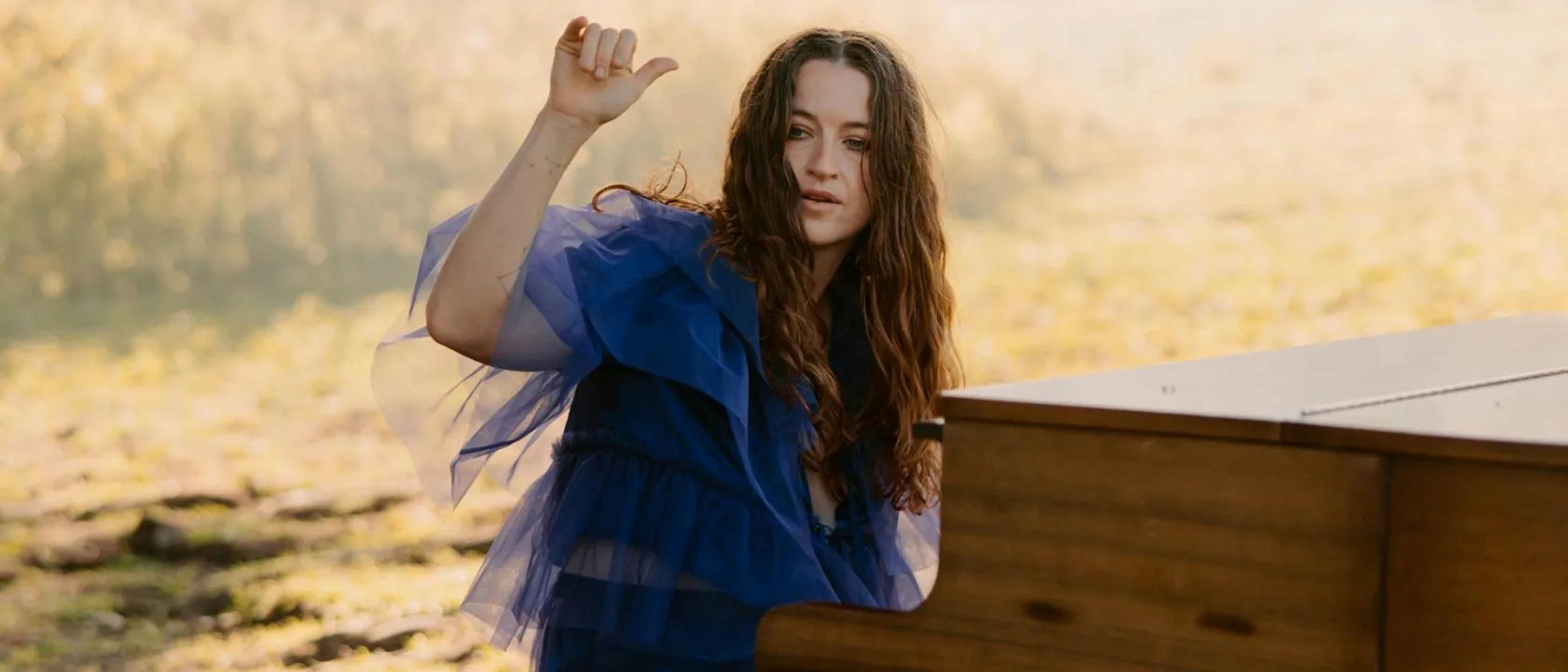 A woman wearing a bue dress sits at a piano, one arm raised