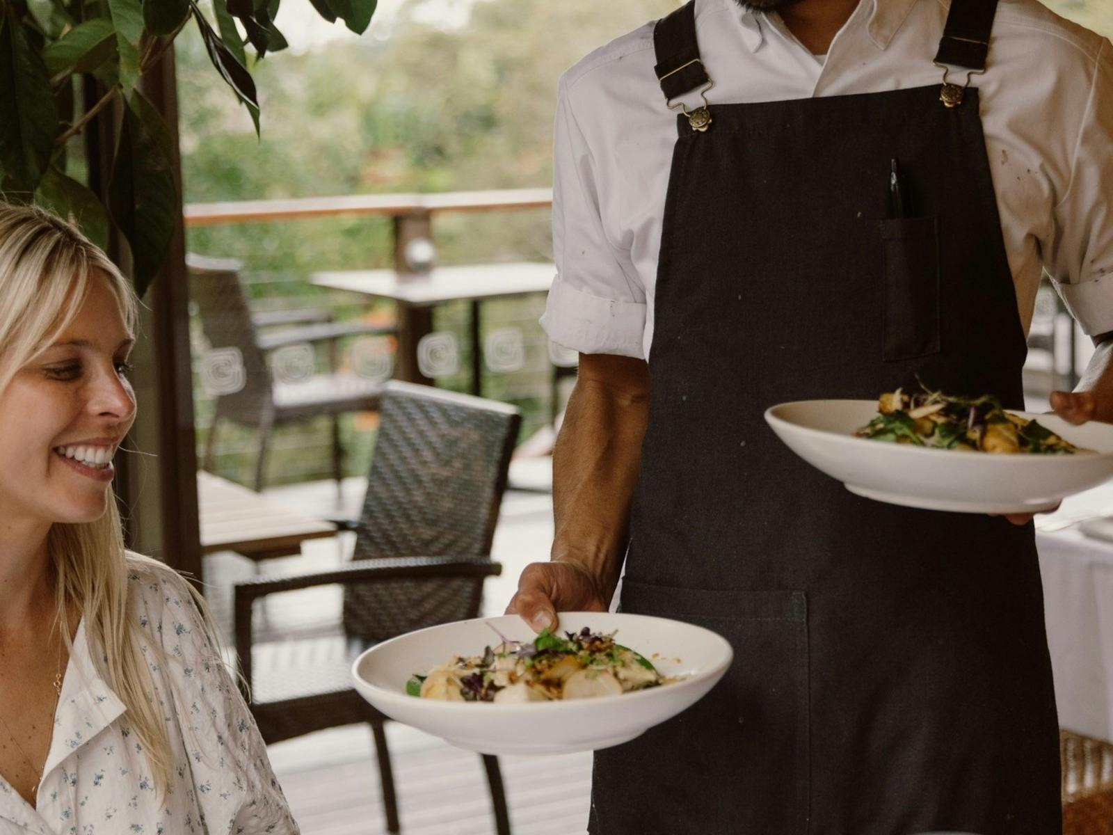 a smiling waiter wearing an apron passing two plates of meals to a waiting smiling woman wearing whi