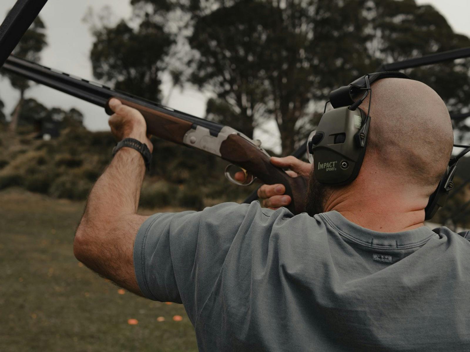 A man in earmuffs hold a shotgun aiming at a clay target.