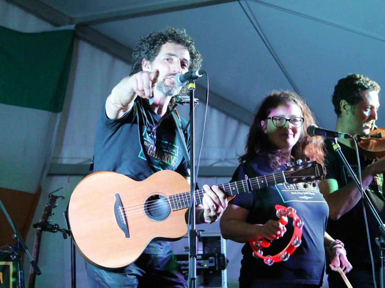 A guitarist points at the camera, with a percussionist looking on and fiddle player in background