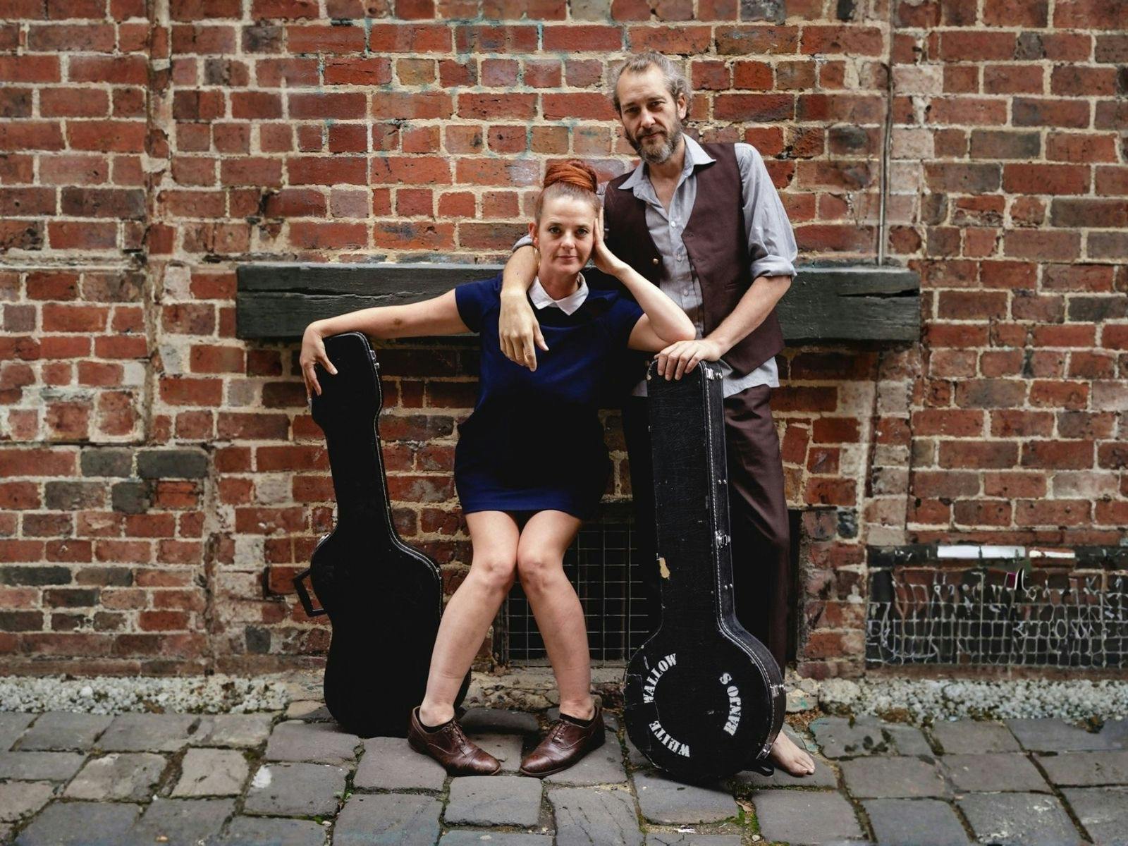 Lizzy & Woody stand with their instruments in front of a brick wall