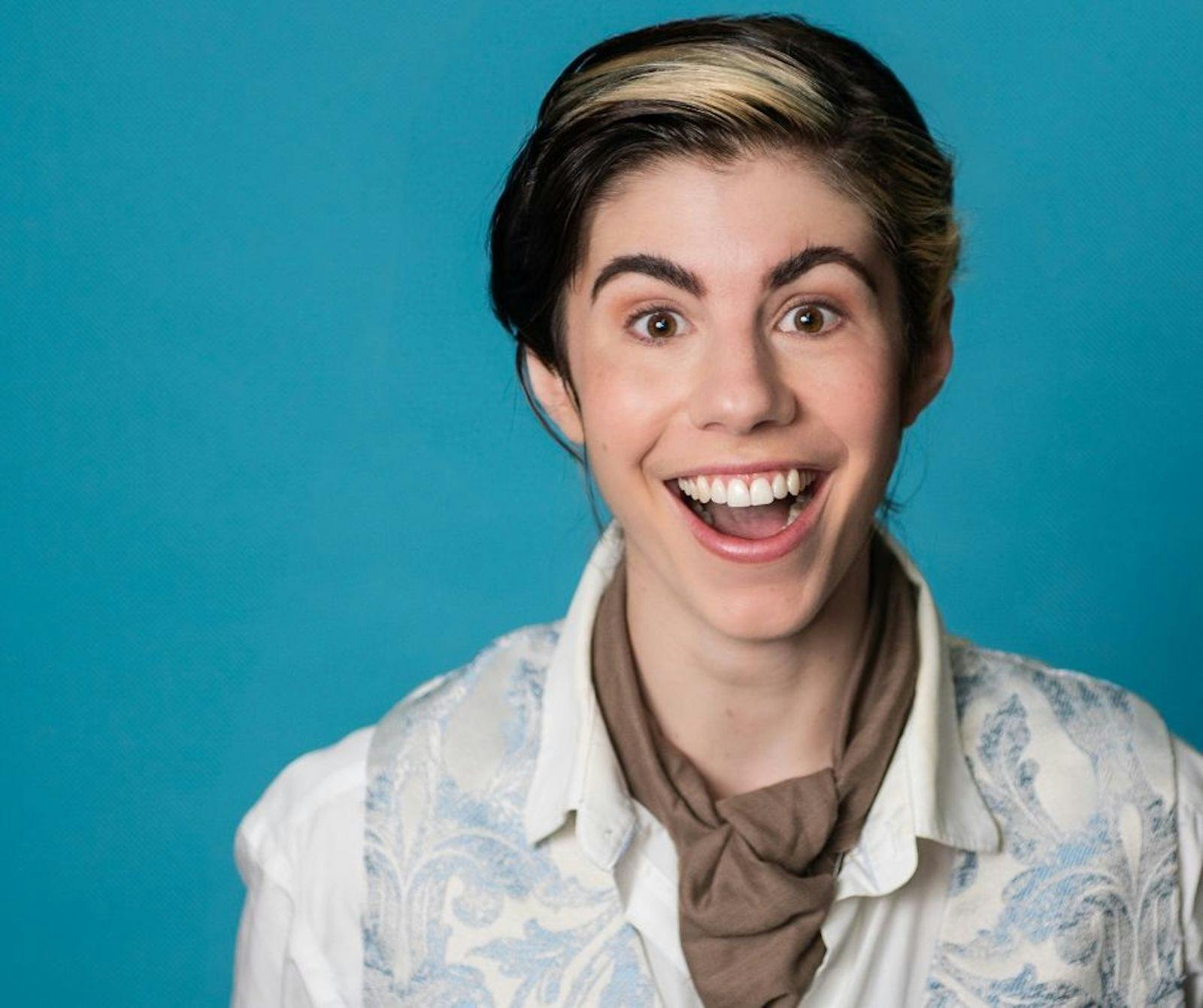 Person smiling in front of blue background, wearing white shirt, blue vest and brown neck scarf.