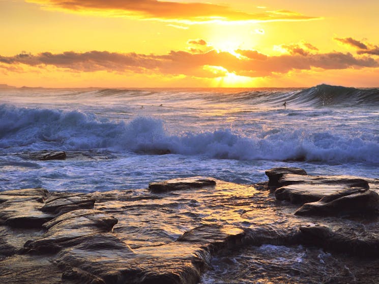 Rocks with waves crashing into them with surfers in background