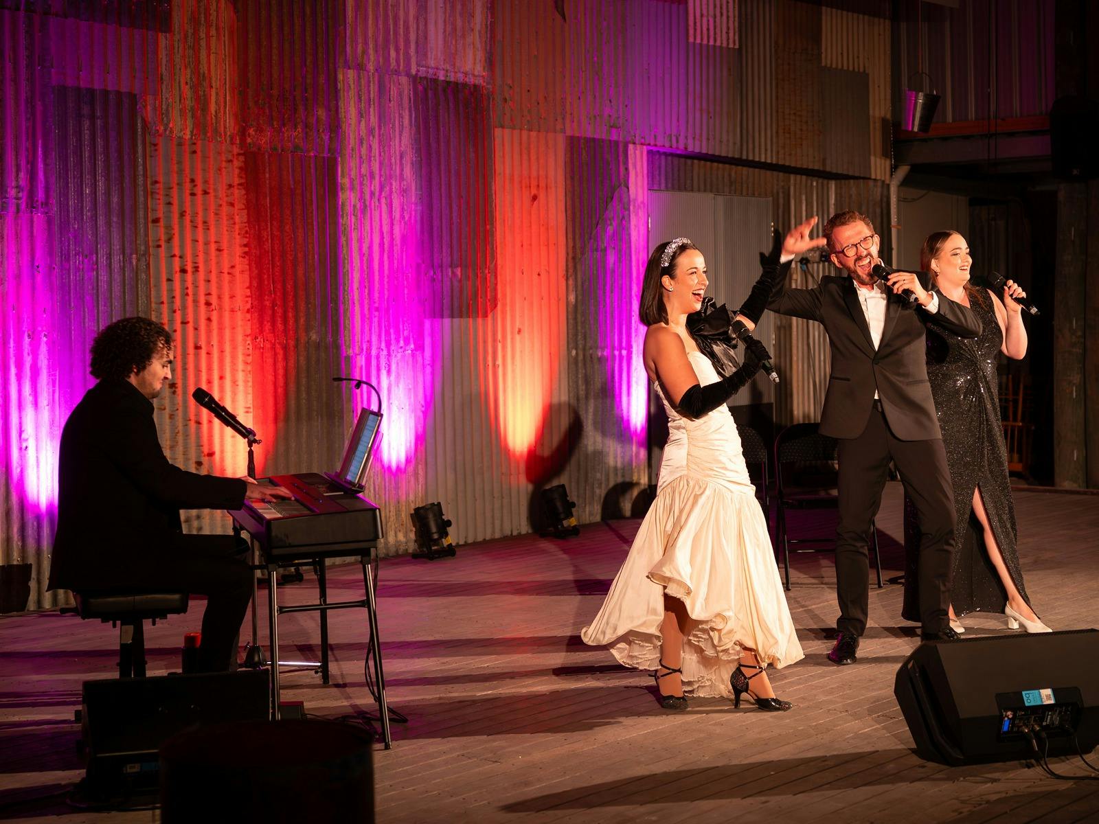 A colourful lit stage with four performers, one playing a keyboard and three standing and singing