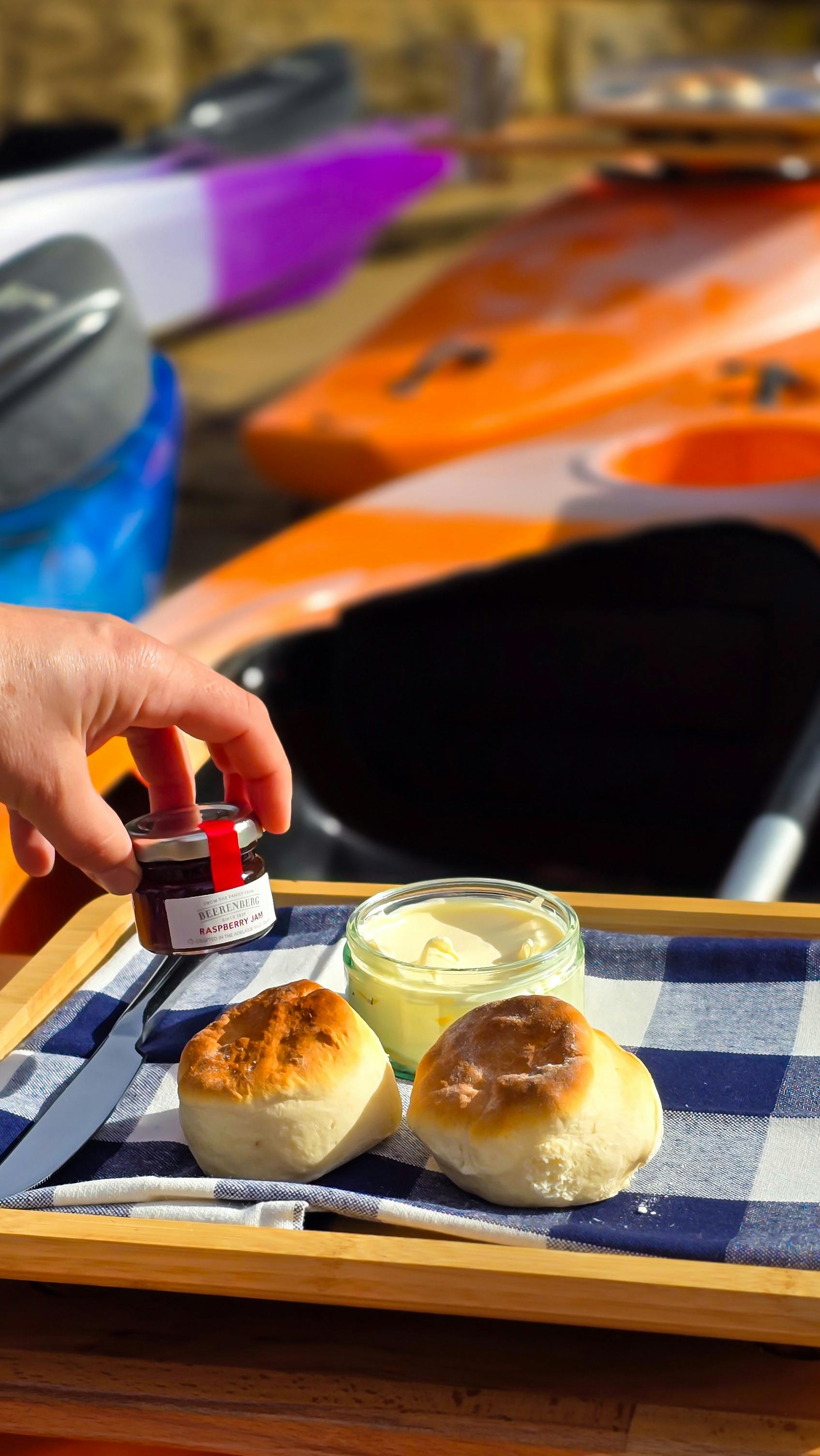 Hand placing down strawberry jam alongside two scones and clotted cream on a wooden board on a kayak