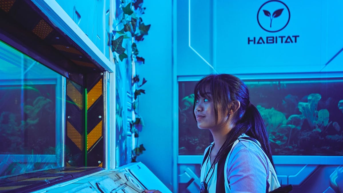 A young woman in a Saboteurs vest works at a control panel in a blue-lit Habitat room.
