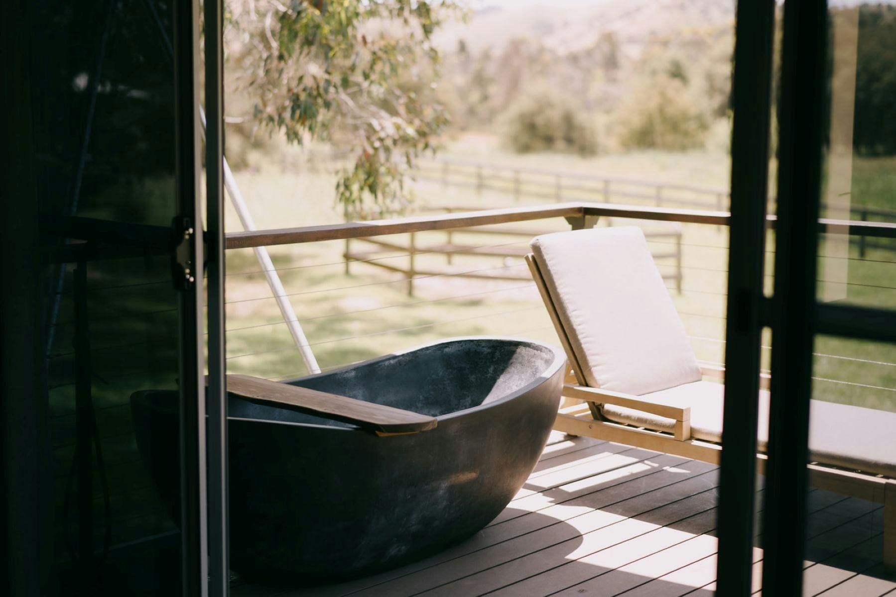A black outdoor bathtub and cushioned lounge chair on a wooden deck overlooking a green landscape.