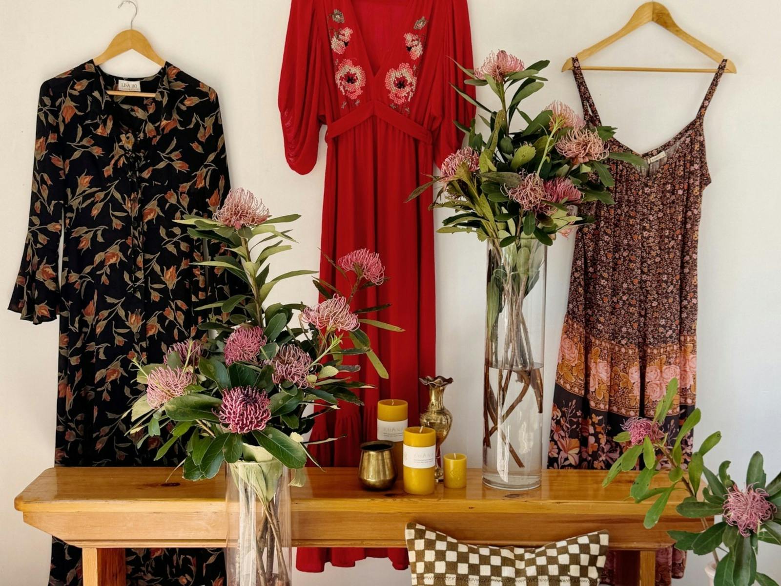 3 bright coloured dresses hanging on a white wall with a timber table with vases of flowers in front