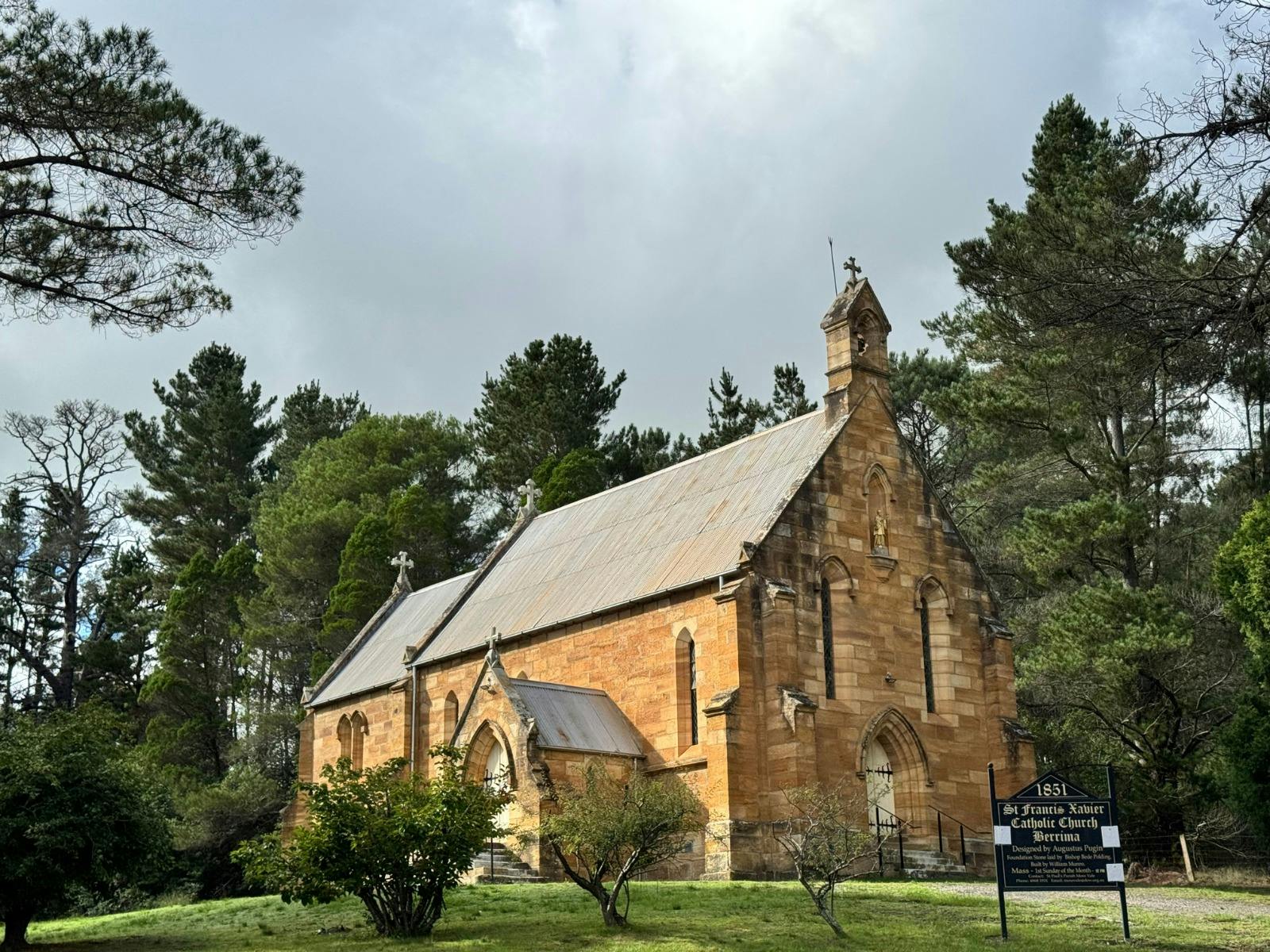 An old church with a tin roof sits among trees. Sun is shining on one side of it.
