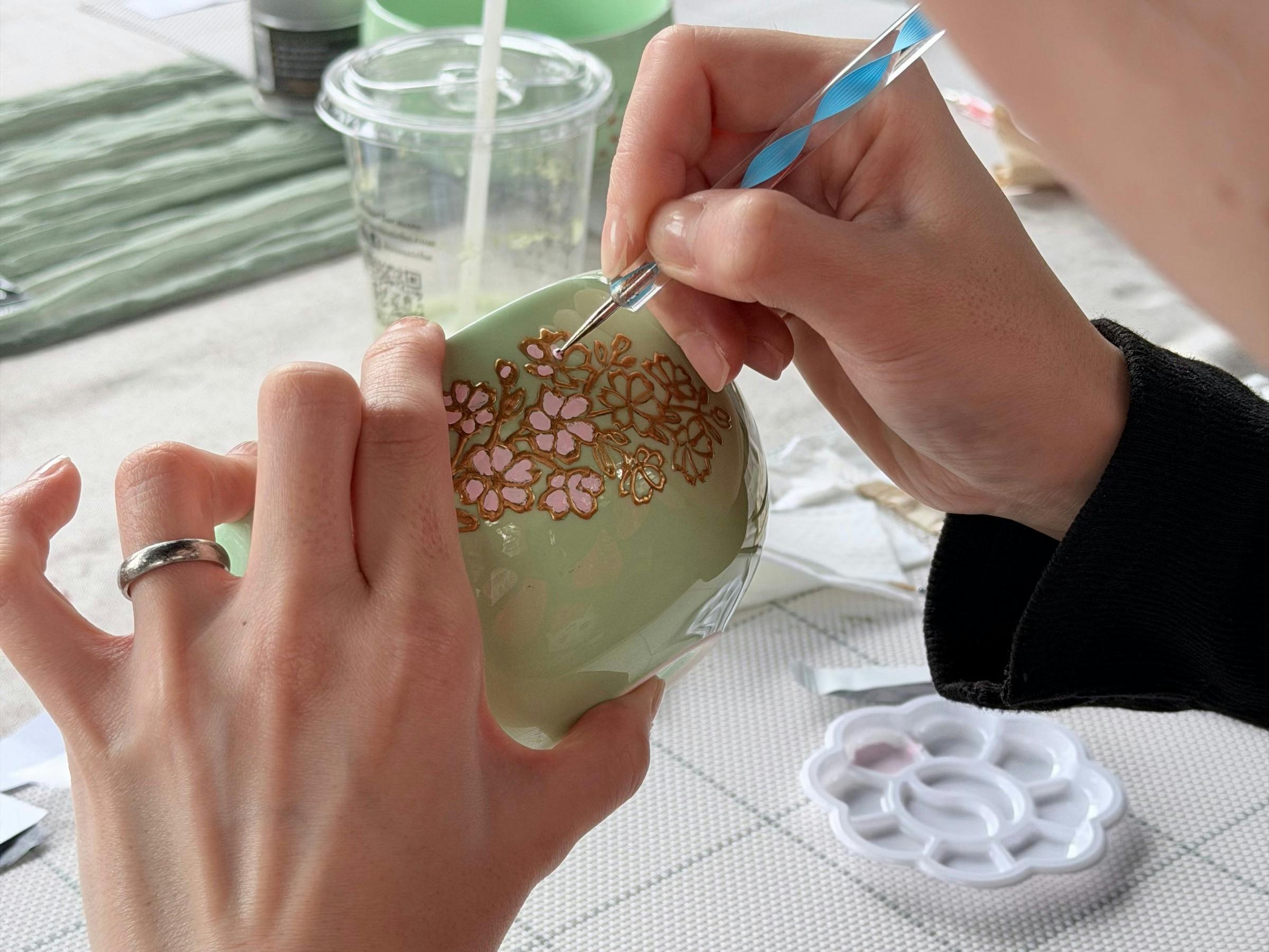 Close-up of hands painting pink and gold flowers on a green ceramic matcha bowl