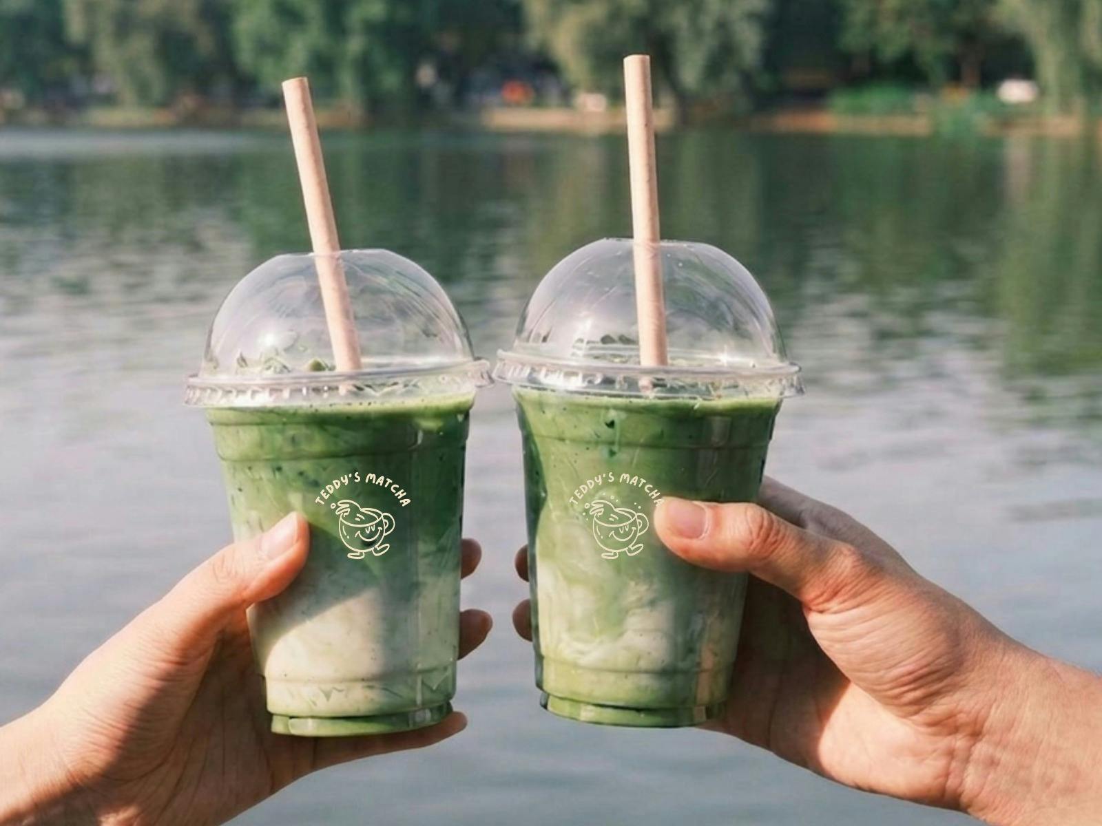 two matcha cups being held in two different hands doing a cheers in front of a lake background