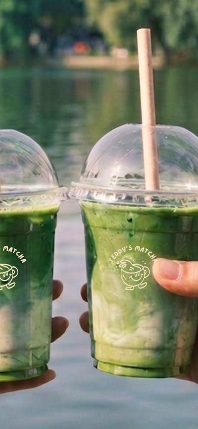 two matcha cups being held in two different hands doing a cheers in front of a lake background