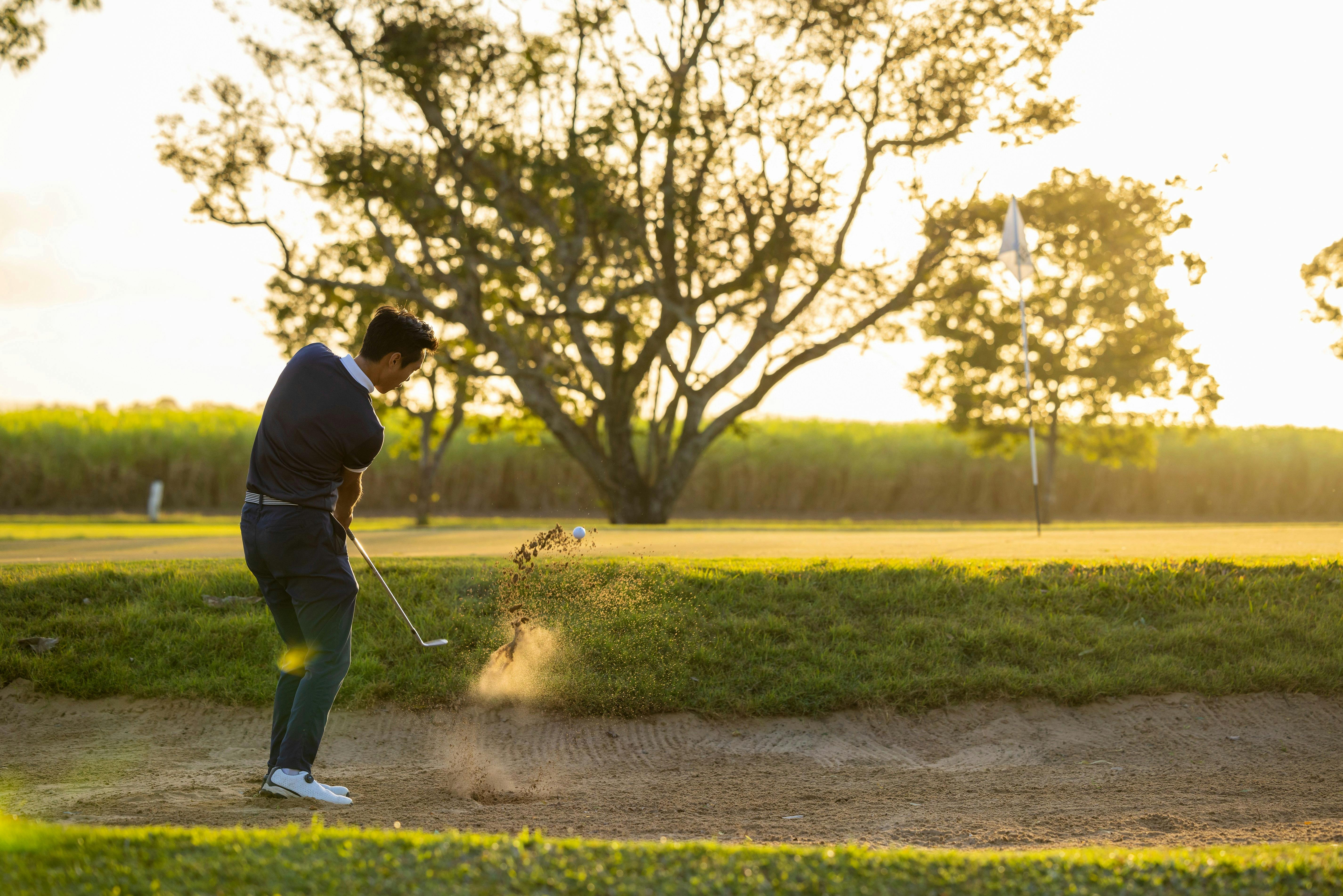 A golfer taking a shot from a sand bunker towards the green with a cane field in the background
