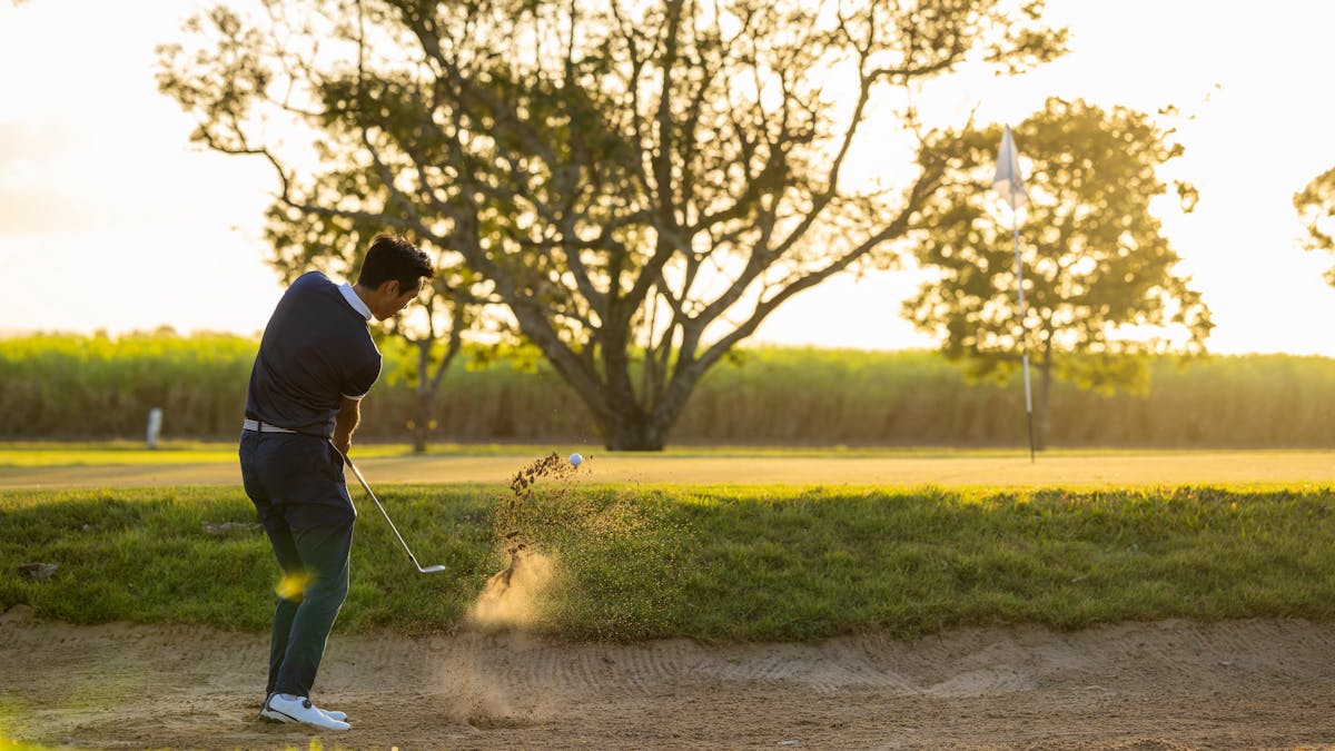 A golfer taking a shot from a sand bunker towards the green with a cane field in the background