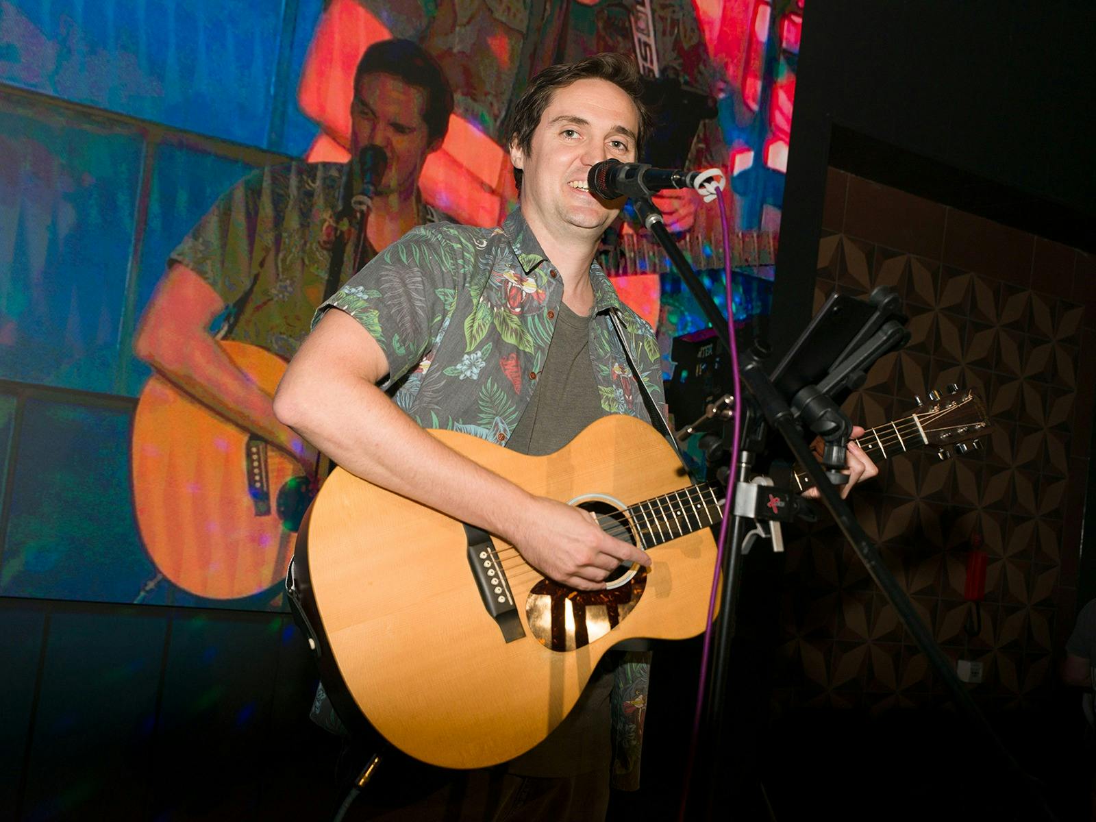Man on a stage singing into a microphone and playing an acoustic guitar