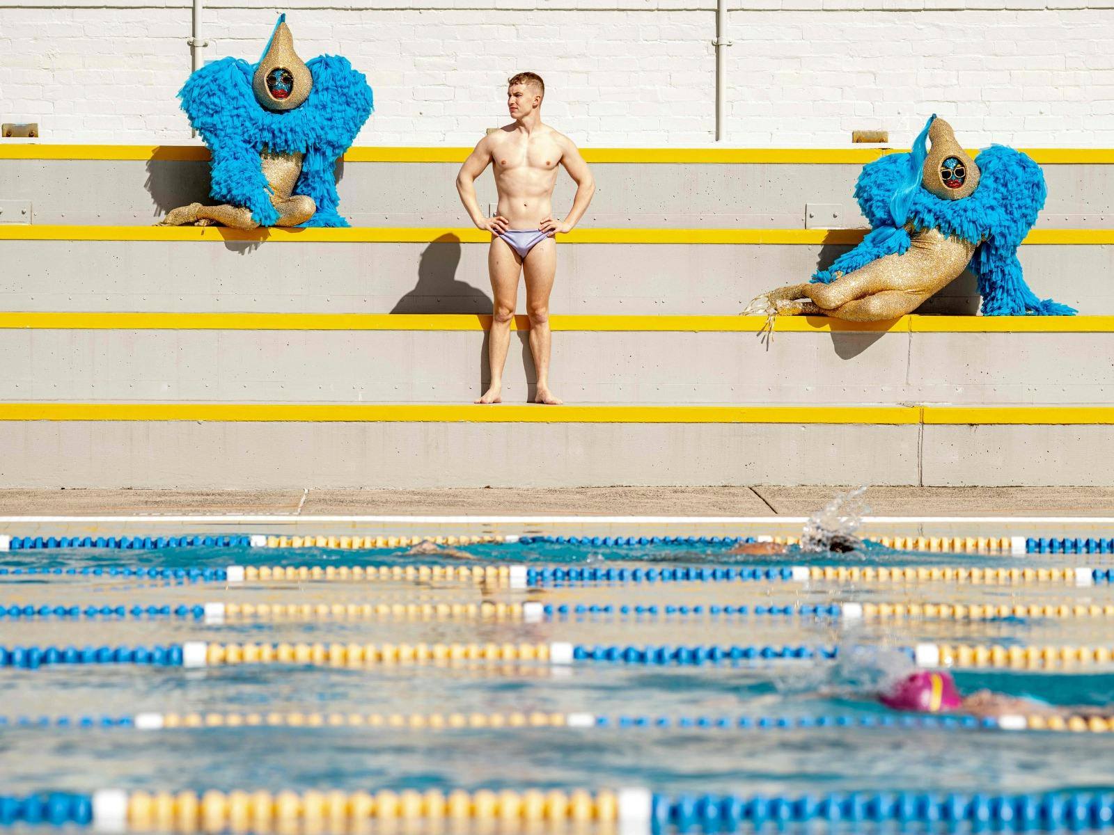 Swimmer and costumed performers pose beside a pool in a theatrical sporting scene.