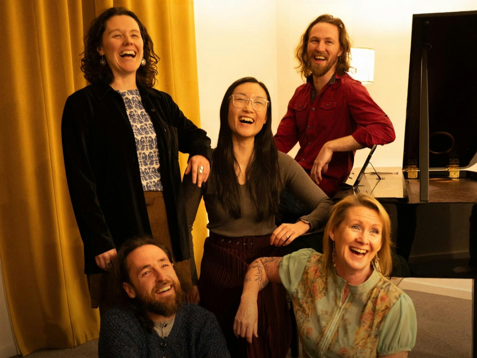 5 performers gathered together laughing next to a grand piano