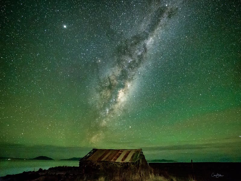 A small dark shed on a hill against a backdrop of a green starry night sky.