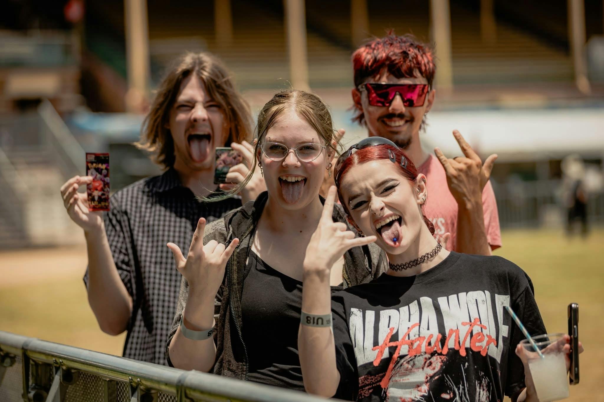 Four young festival attendees making rock hand signs and cheering with tongues out, festival grandst