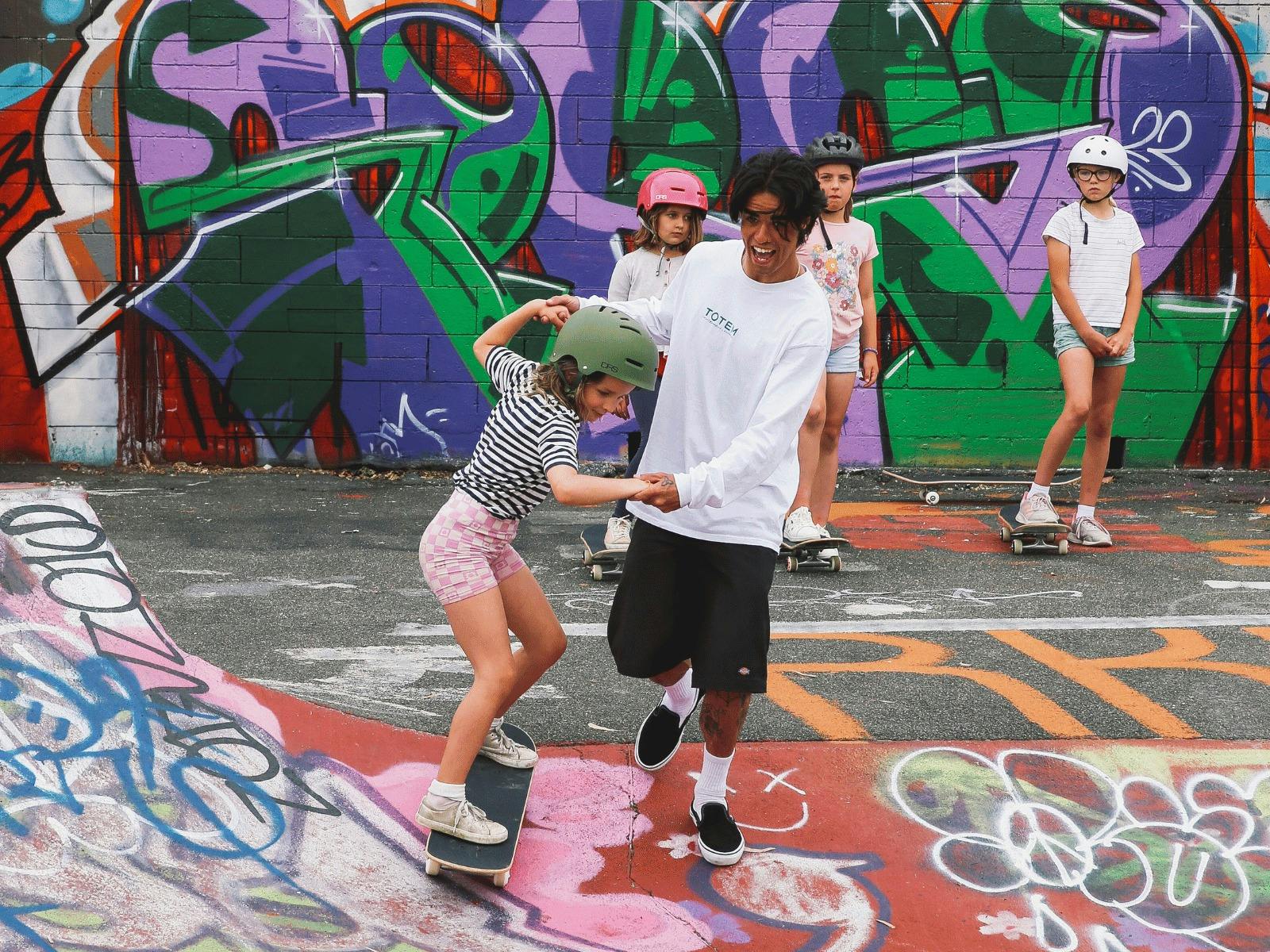young girl on skateboard being helped by young man in front of grafitti wall