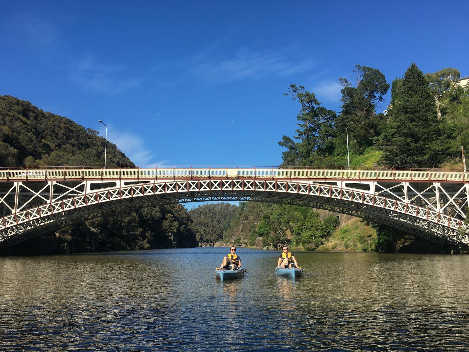 Two people in two blue pedal power kayaks paddle under an arched iron work bridge in the background