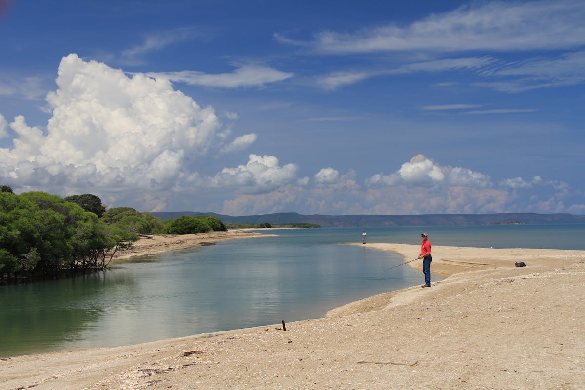 Ma stands fishing in shallows with mangroves on shore, under blue skies.