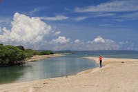 Ma stands fishing in shallows with mangroves on shore, under blue skies.