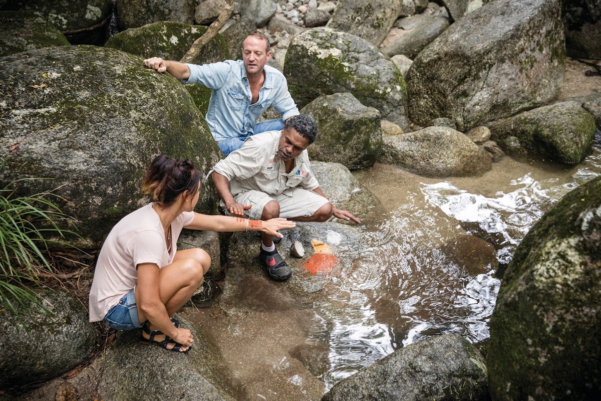 Aboriginal guide at Mossman Gorge with guests, demonstrating ochre paints by the creek.