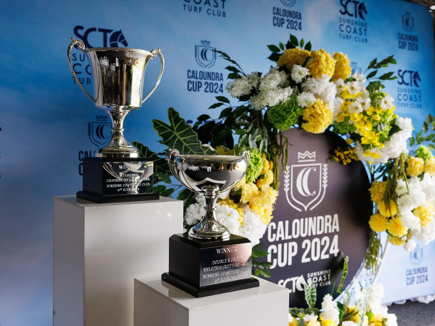 two large silver trophies on plinths in front of a blue media wall and circular floral arrangement