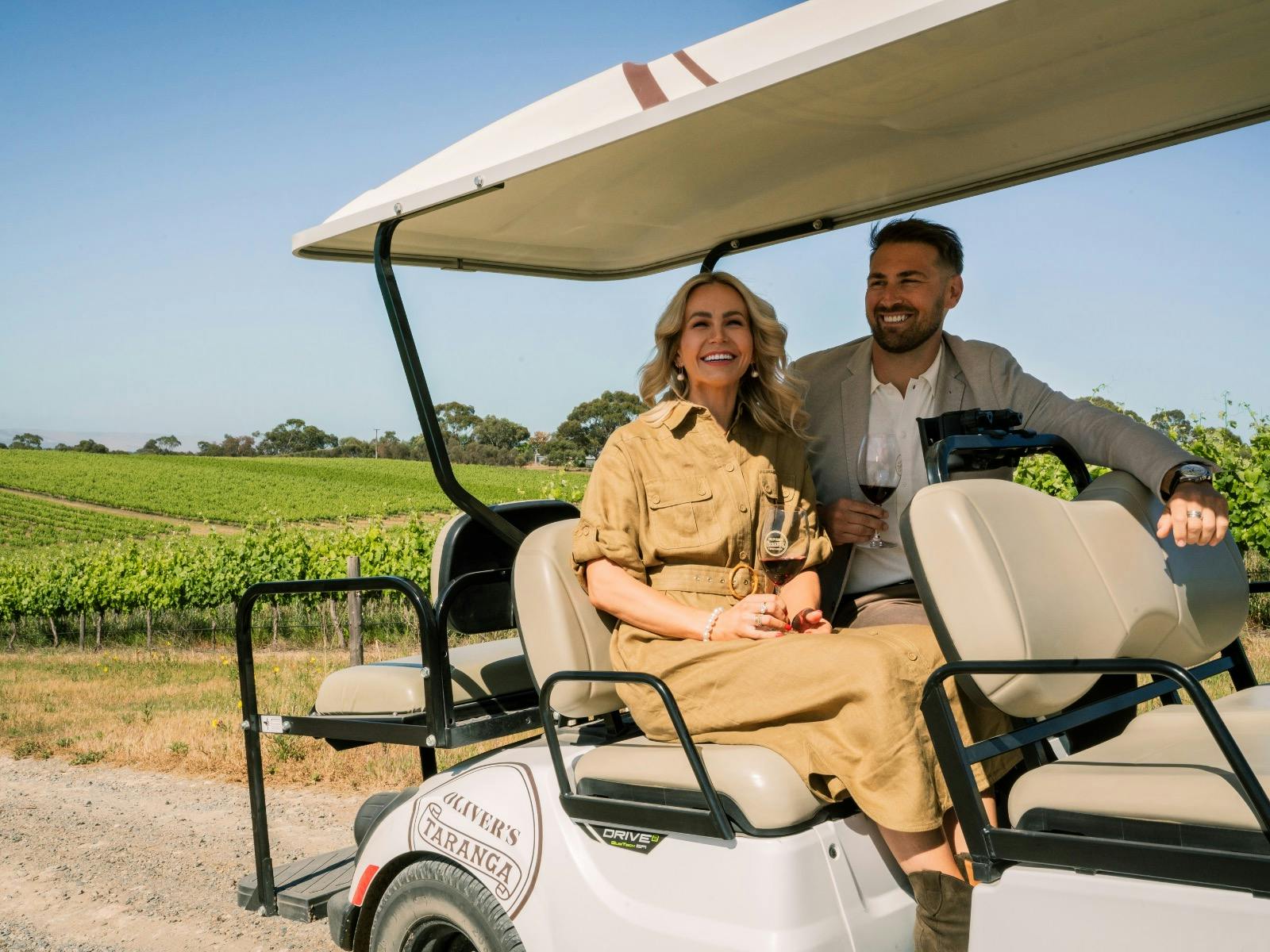 A smiling happy couple sit in the rear seats of a luxury golf buggy in the vineyard.
