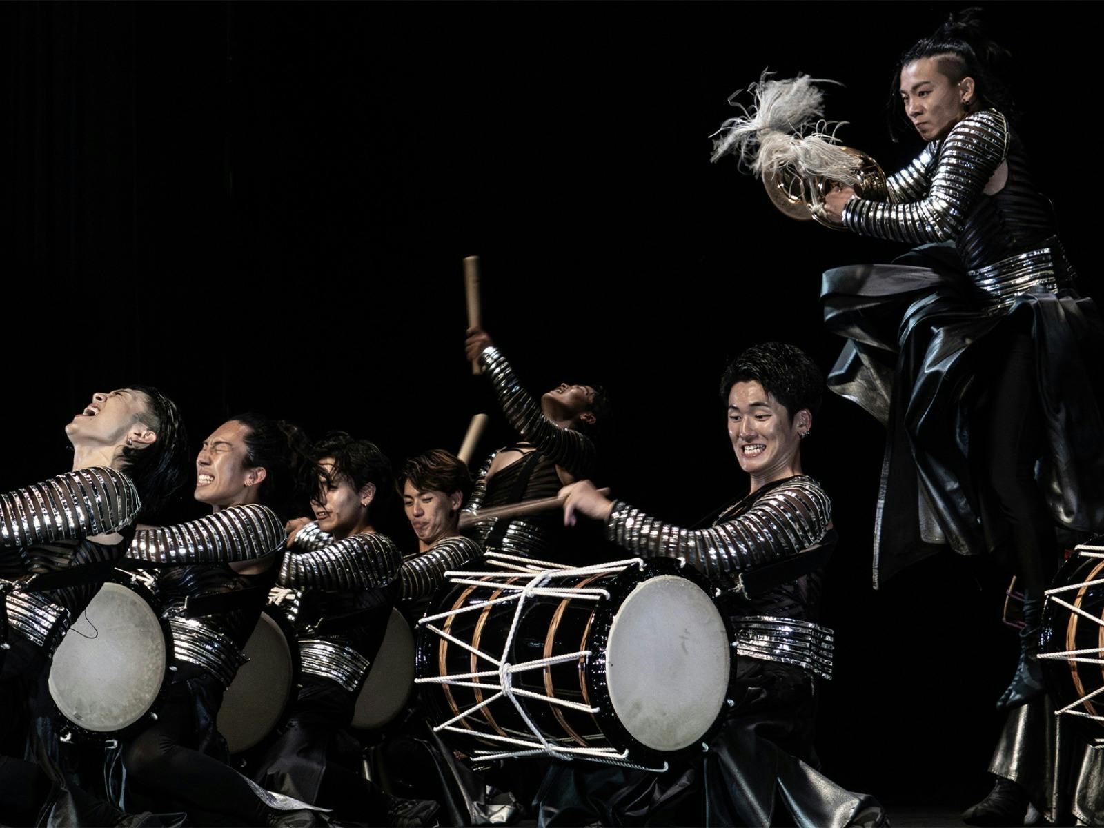 Energetic performers in metallic silver costumes play taiko drums on stage while one leaps mid-air.