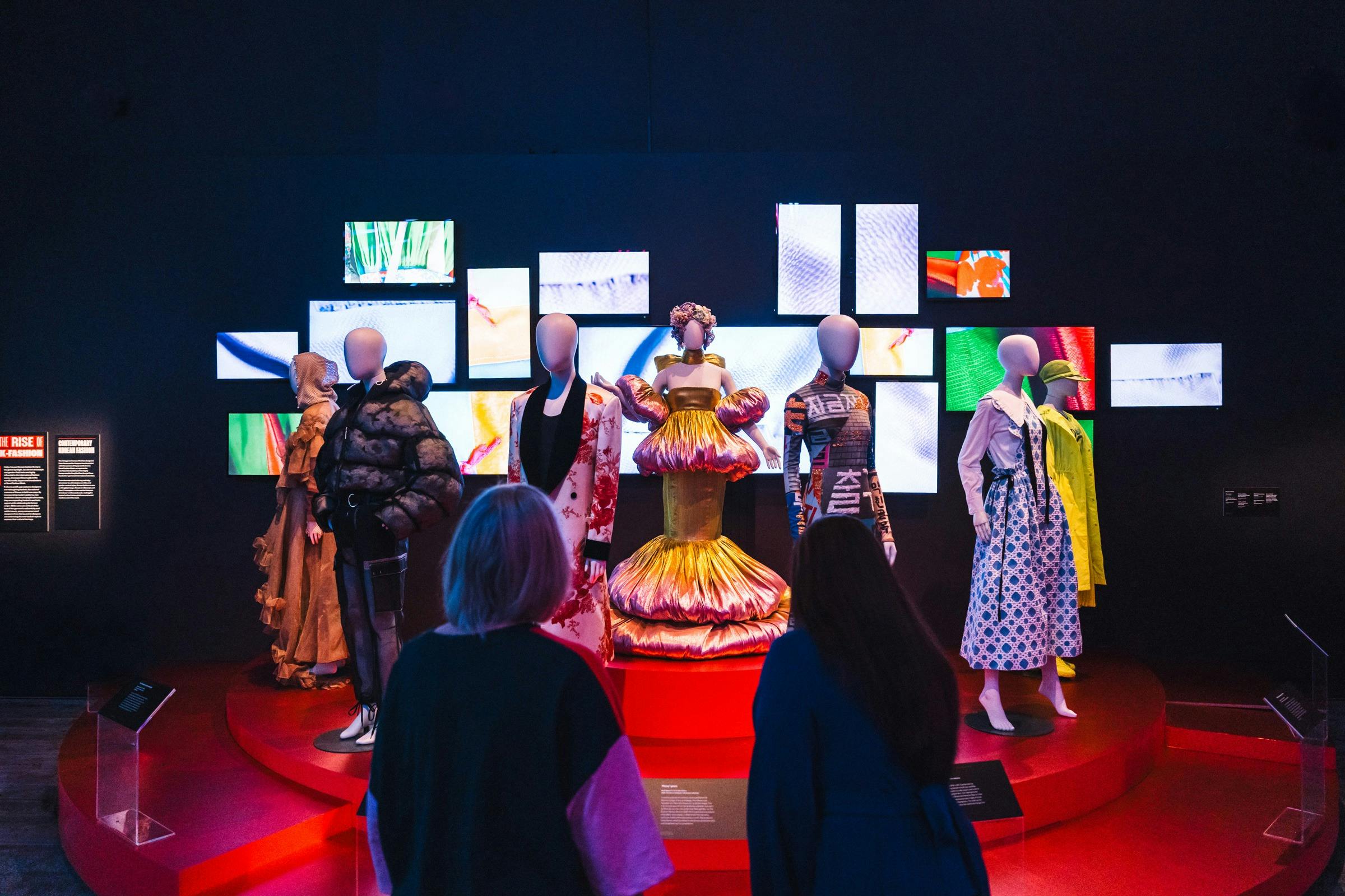 A dimly lit exhibit showcases mannequins dressed in Korean fashion on a red circular platform.