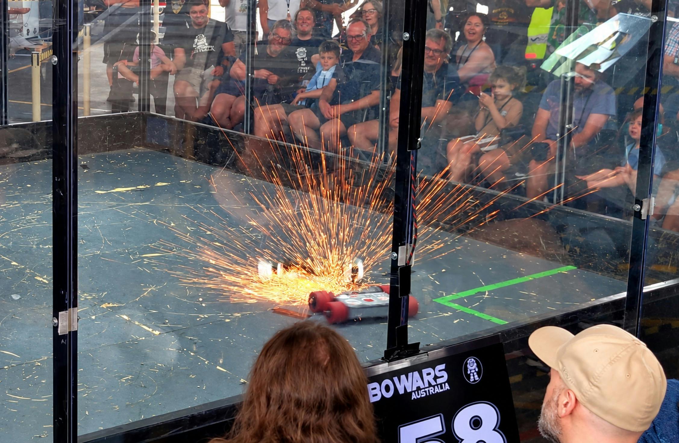 Combat robots clash in a shower of sparks as spectators watch from outside the enclosed arena.