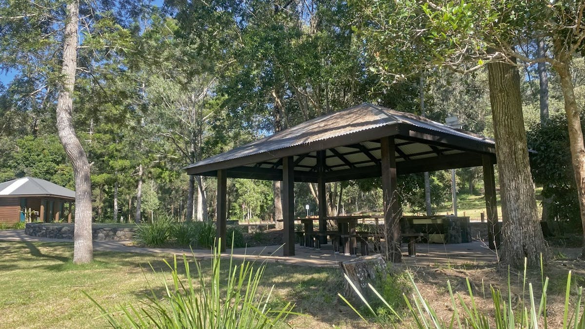 Sheltered picnic tables in grassy area with a toilet block in the background.