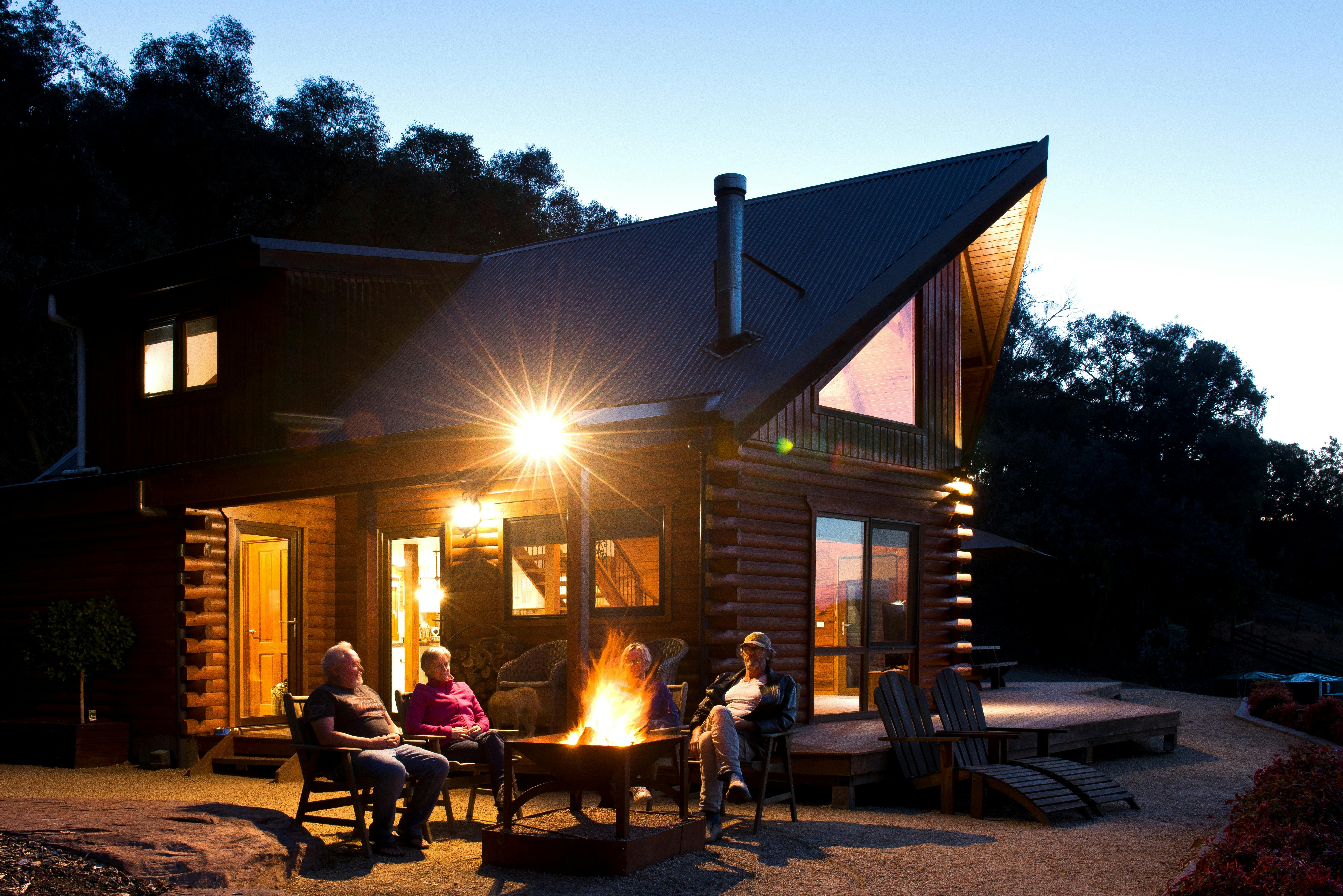 Four guests sit around a glowing fire pit in front of a timber log cabin at dusk in the High Country