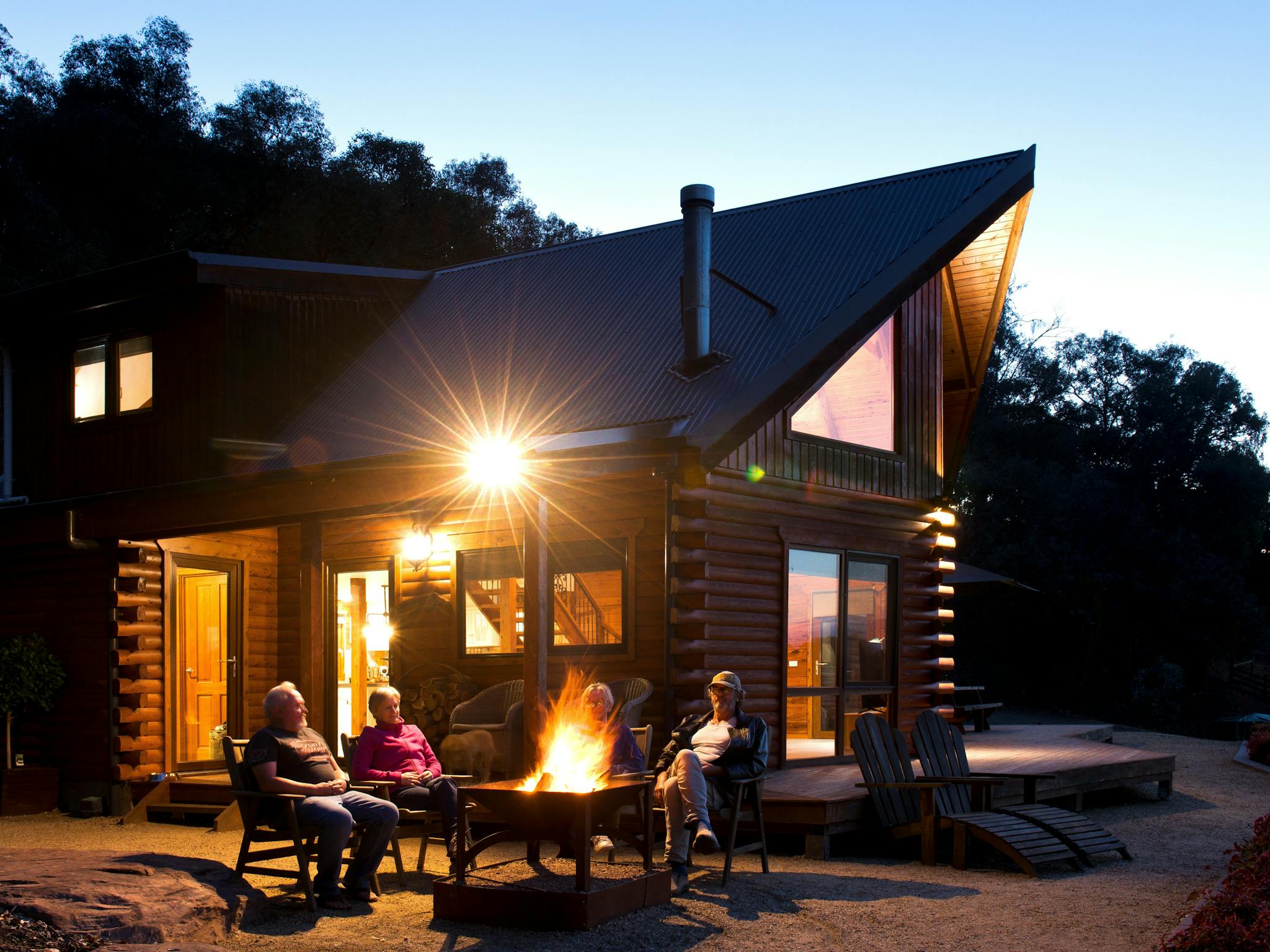 Four guests sit around a glowing fire pit in front of a timber log cabin at dusk in the High Country