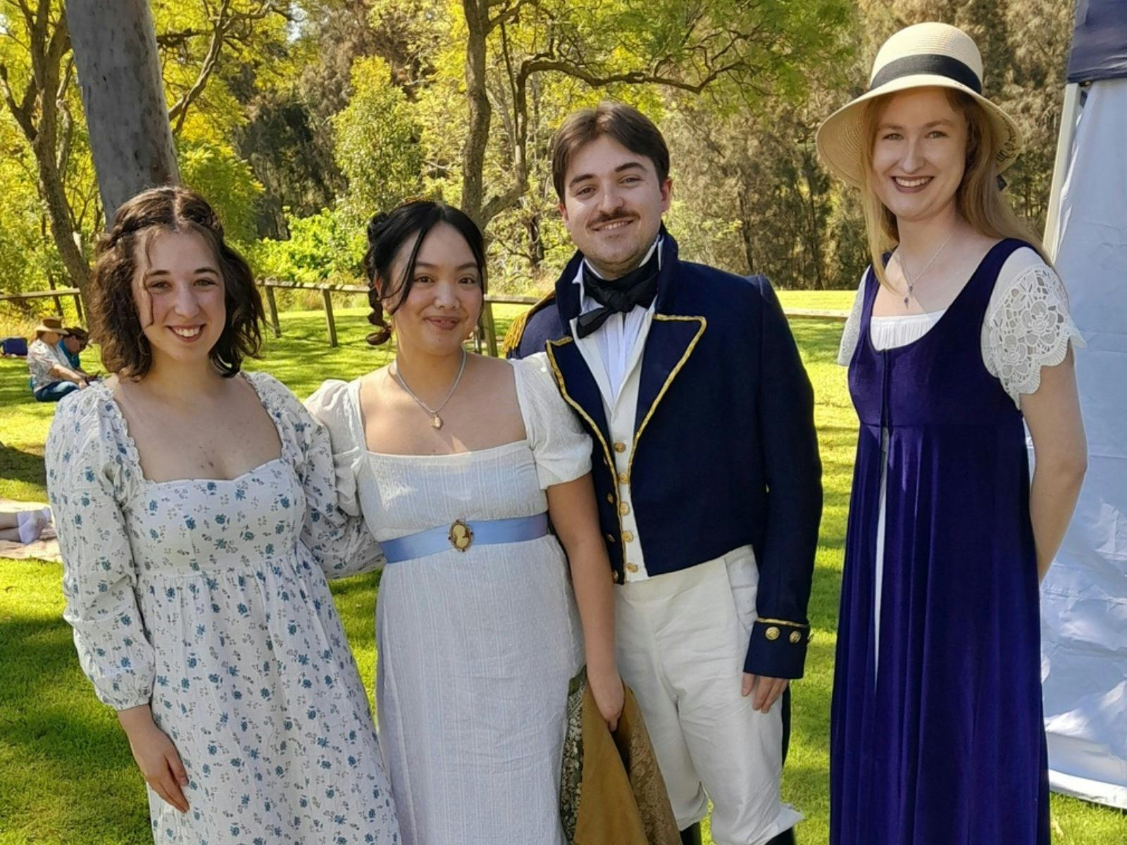 Four young people dressed in Regency clothes stand under sun lit trees