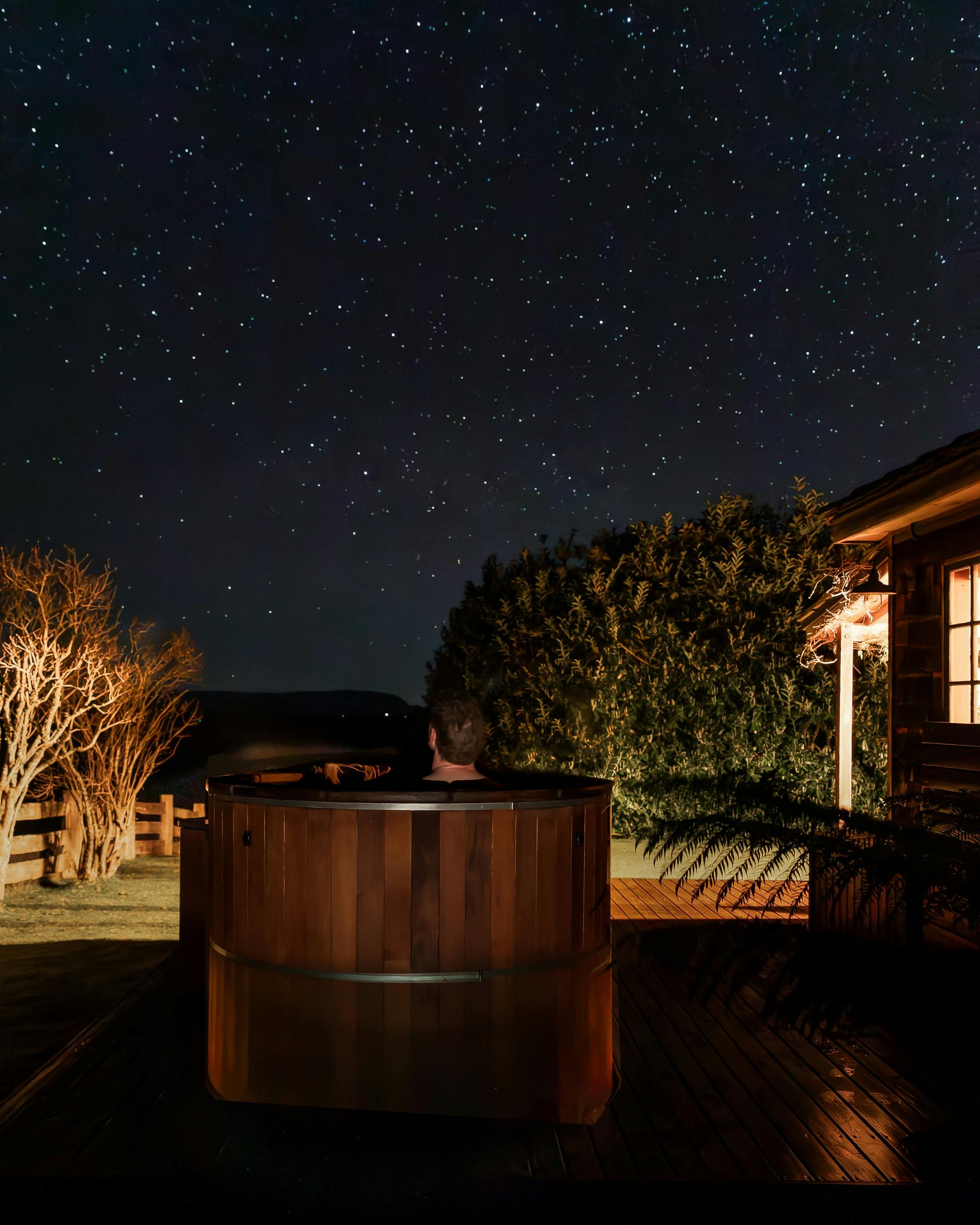A person relaxes in a cedar hot tub at night, gazing up at a clear starry sky.