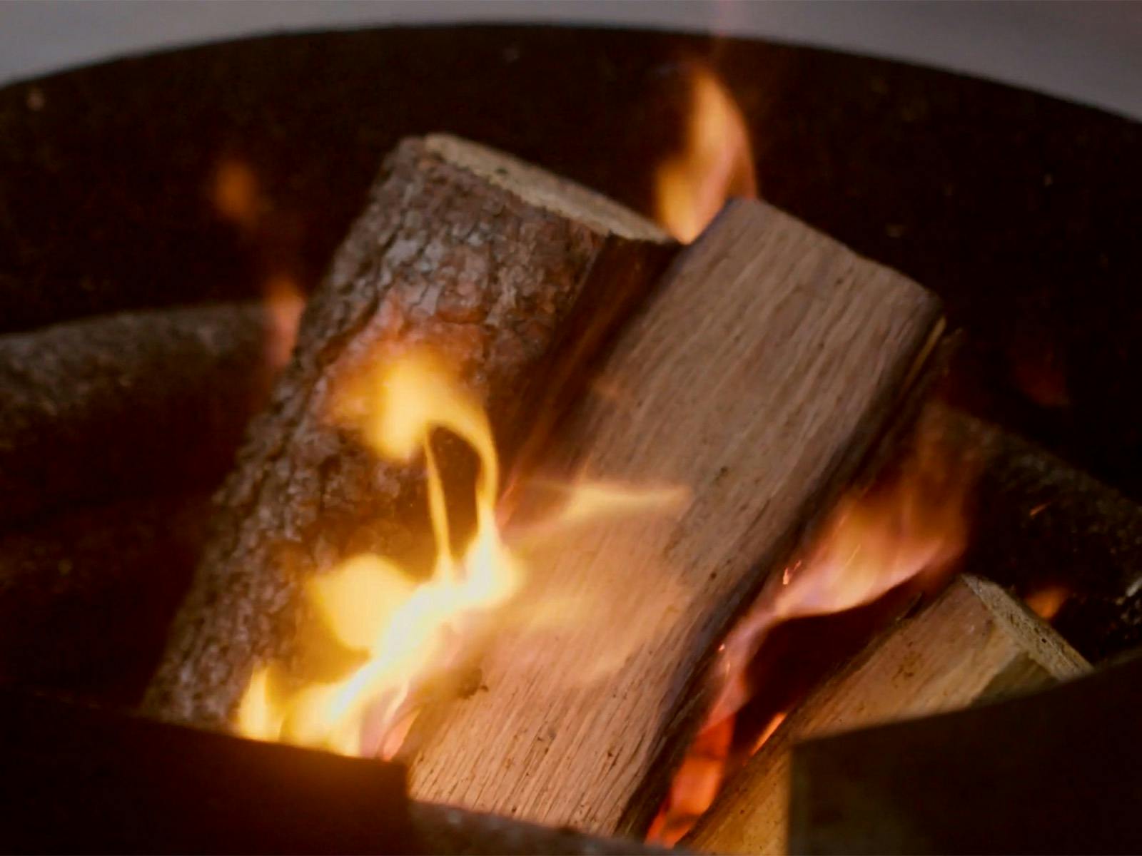 Flames dance brightly against a dark metal firepit, glowing in the low light of the evening