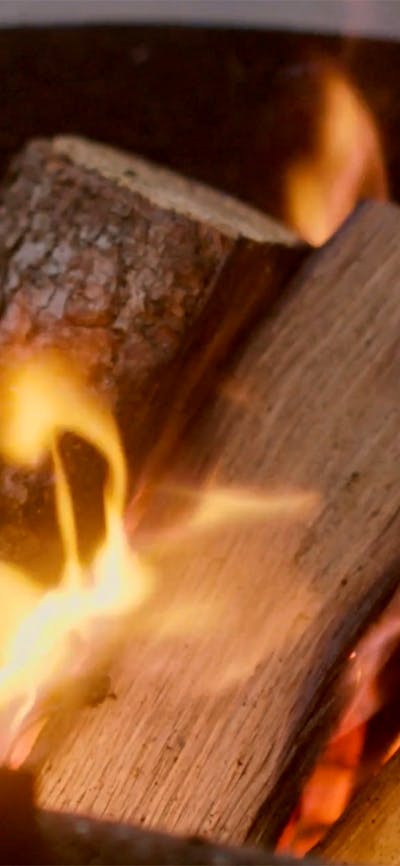 Flames dance brightly against a dark metal firepit, glowing in the low light of the evening