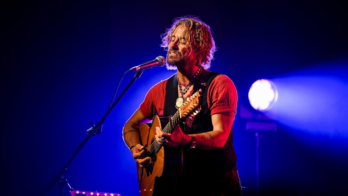 A musician sings and plays acoustic guitar on stage under blue and purple concert lighting.