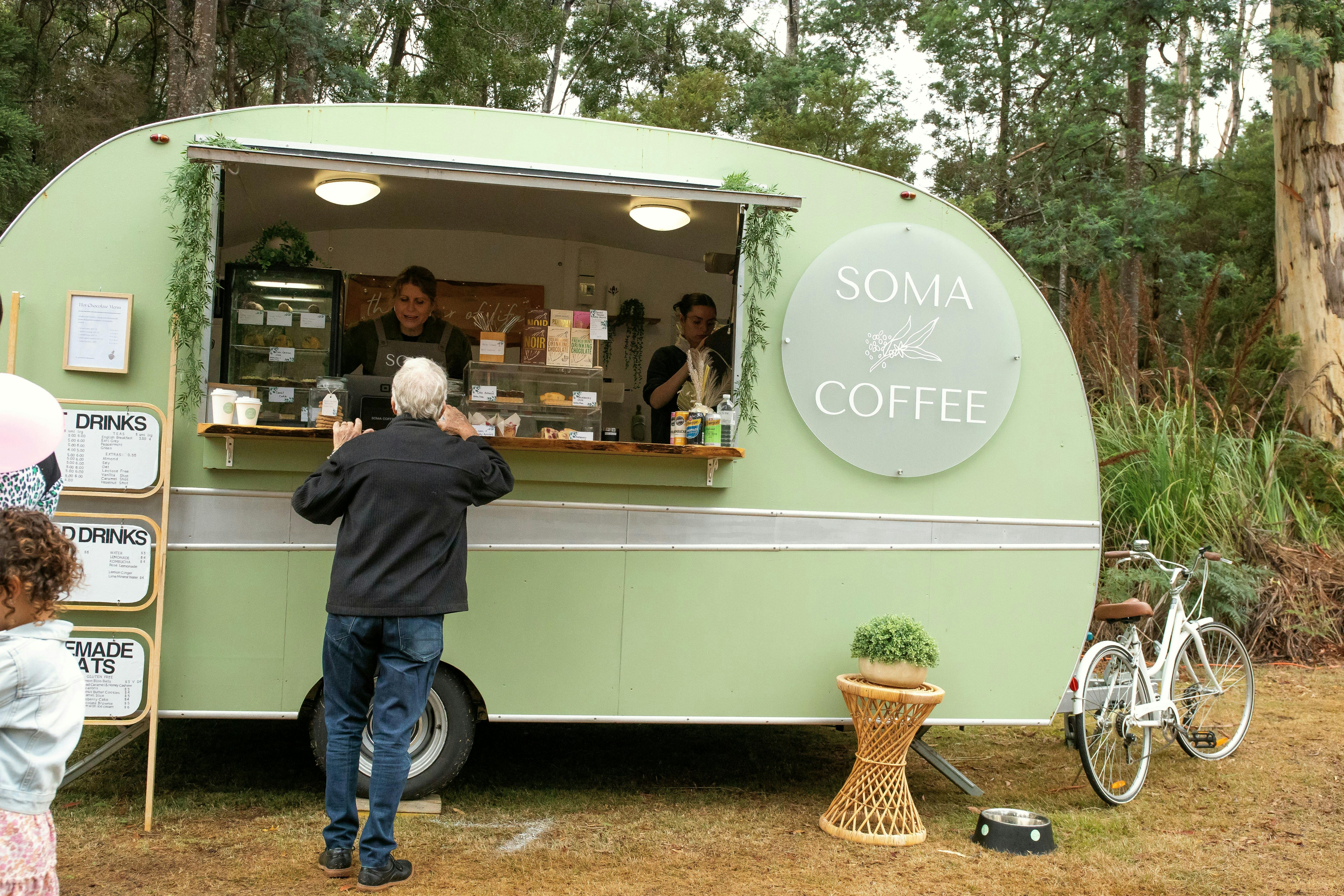 Green vintage coffee van parked in front of native scrub with bike propped at side and a customer