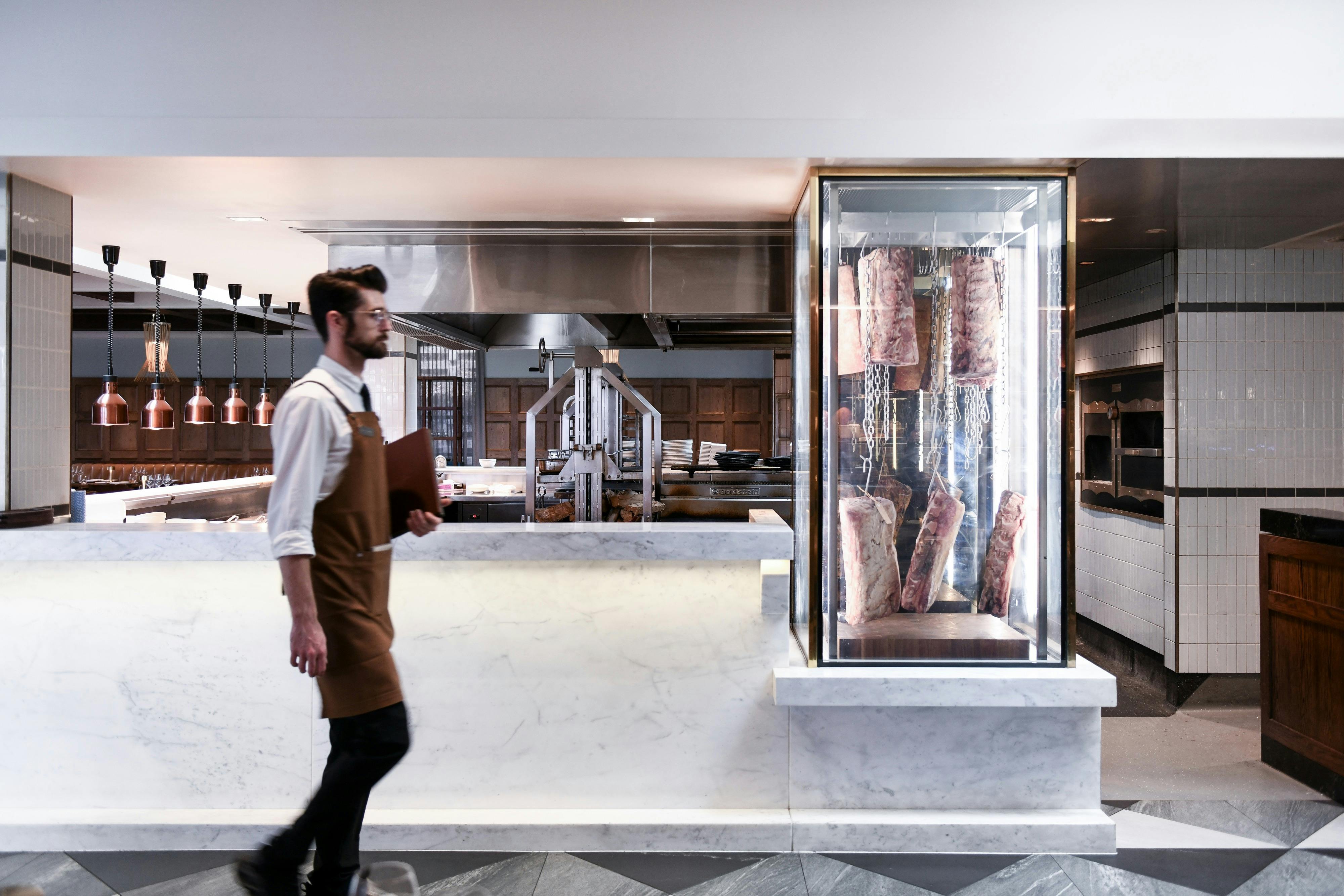 Waiter in apron walks past open kitchen with meat display and marble counter.
