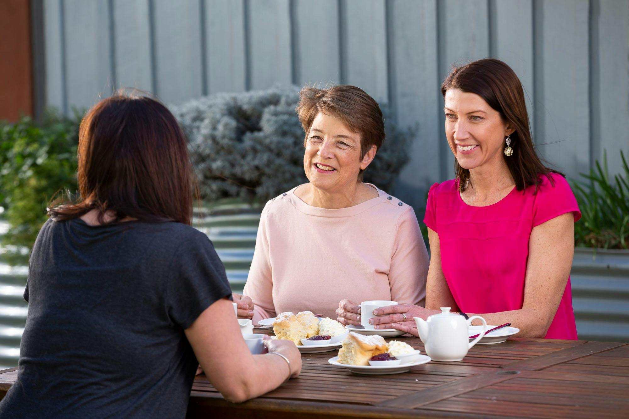 3 Women at an outdoor table chatting whilst eating a sweet breakfast with coffee.